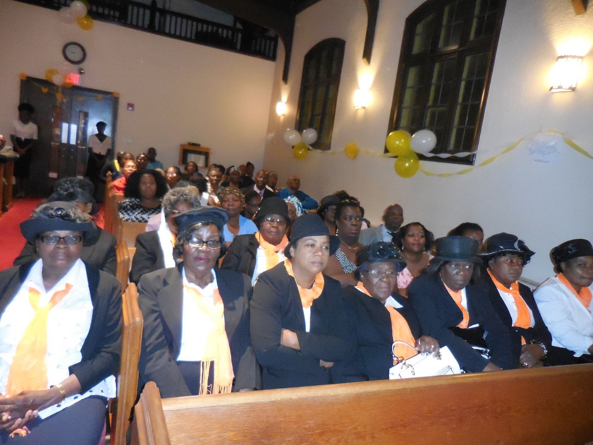 A group of people wearing matching black jackets and yellow scarves sit in church pews during an indoor event.