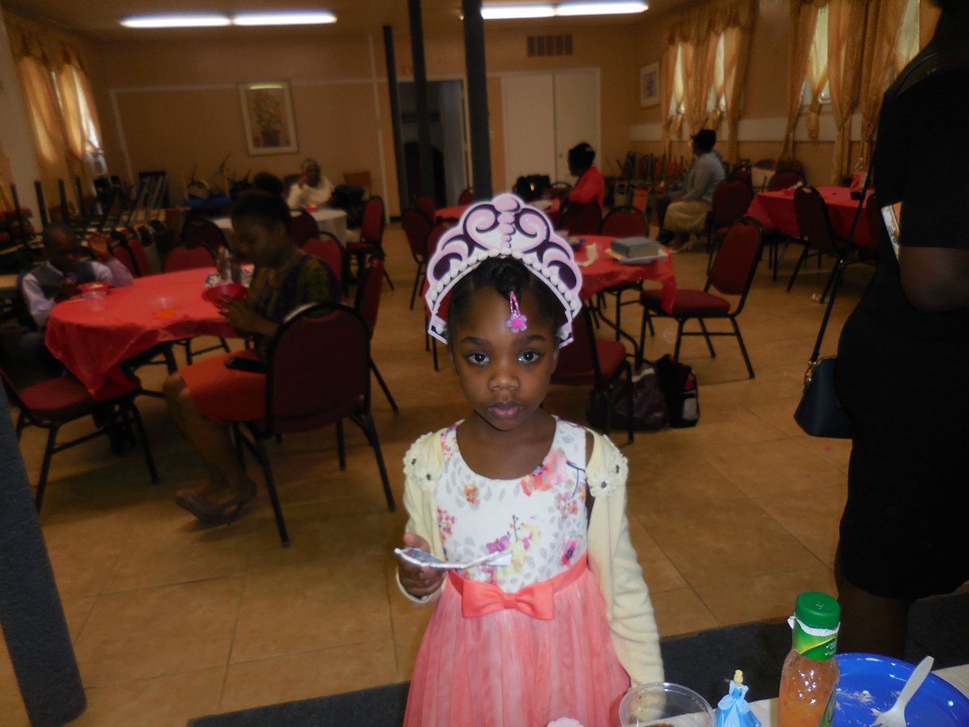 A person wearing a pink floral dress and a decorative crown stands in a room with red tablecloths and chairs.