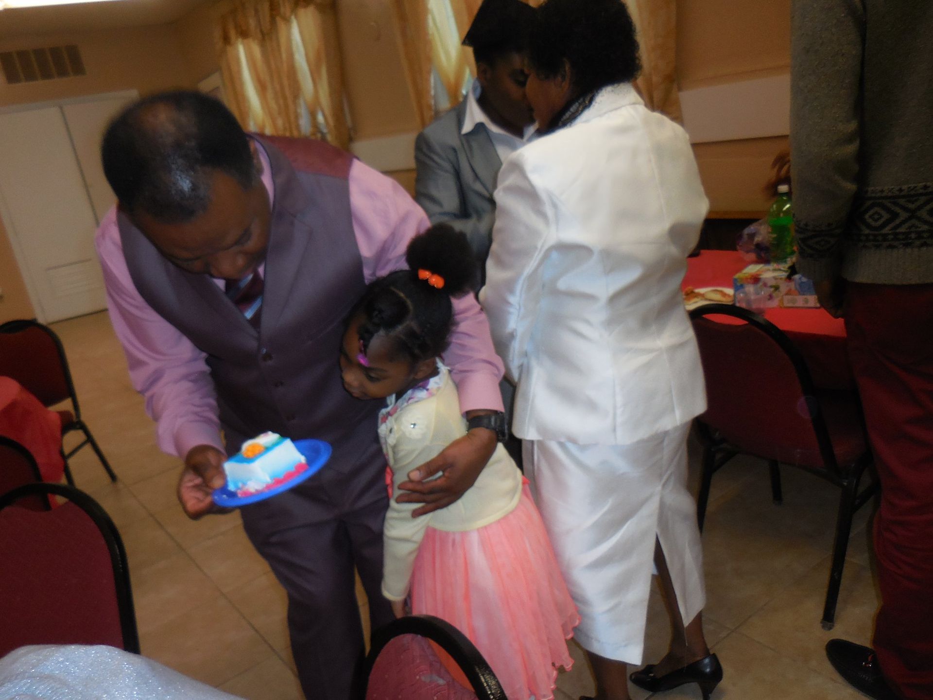 An adult in a vest holds a plate with a cake for a child in a pink skirt while others gather in a banquet hall.