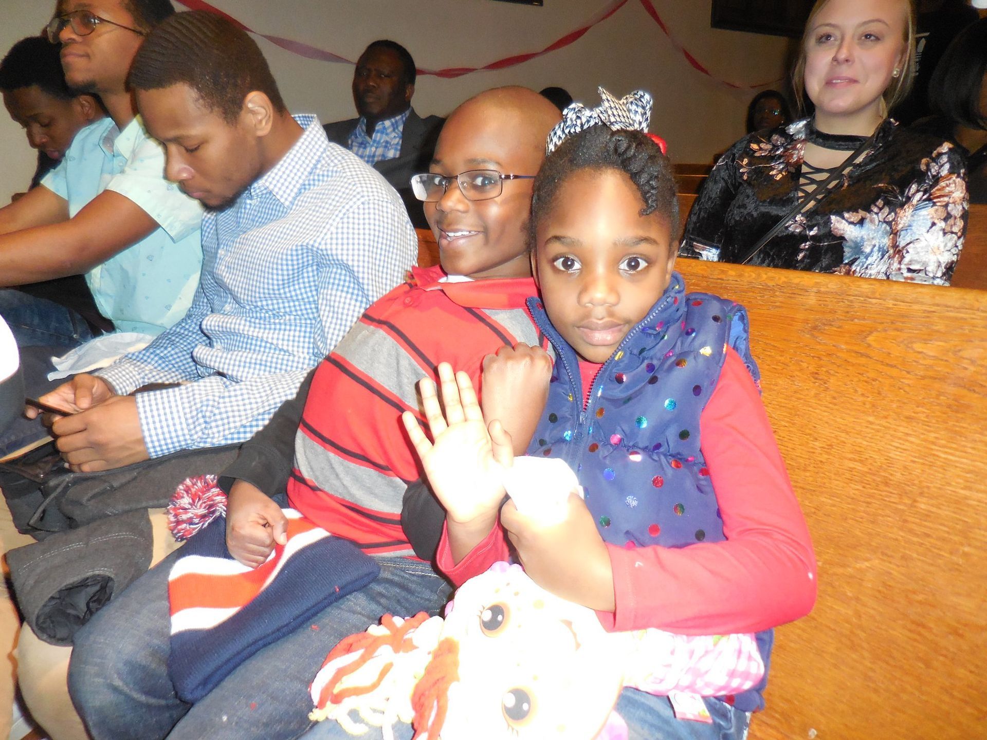 A group of people sitting in church pews, with a girl holding a doll in the foreground.