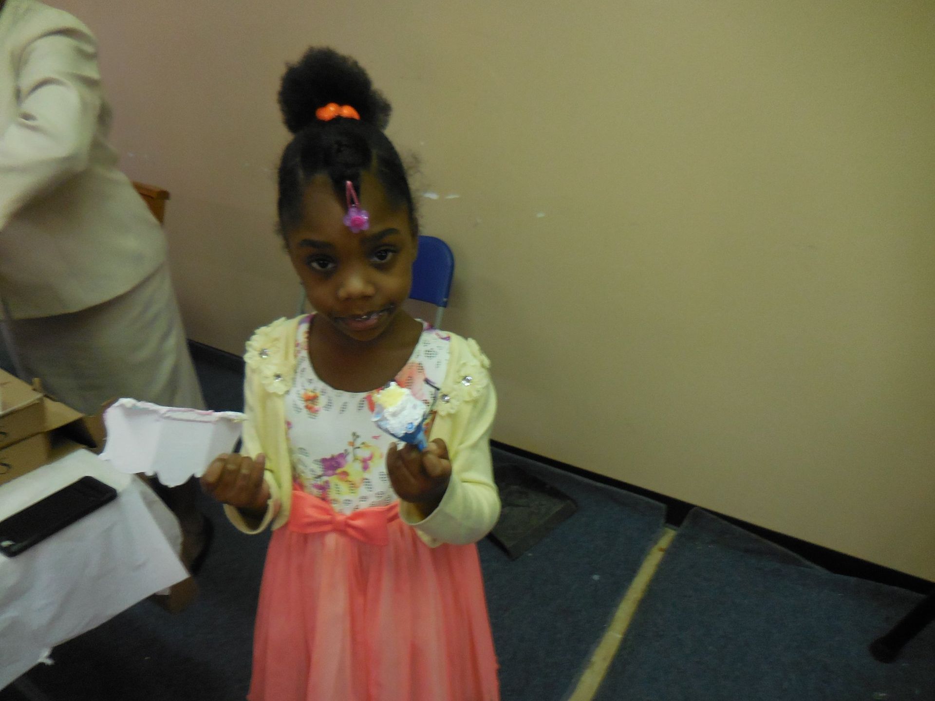 A child wearing a floral dress and yellow cardigan holds a small craft item in an indoor setting.