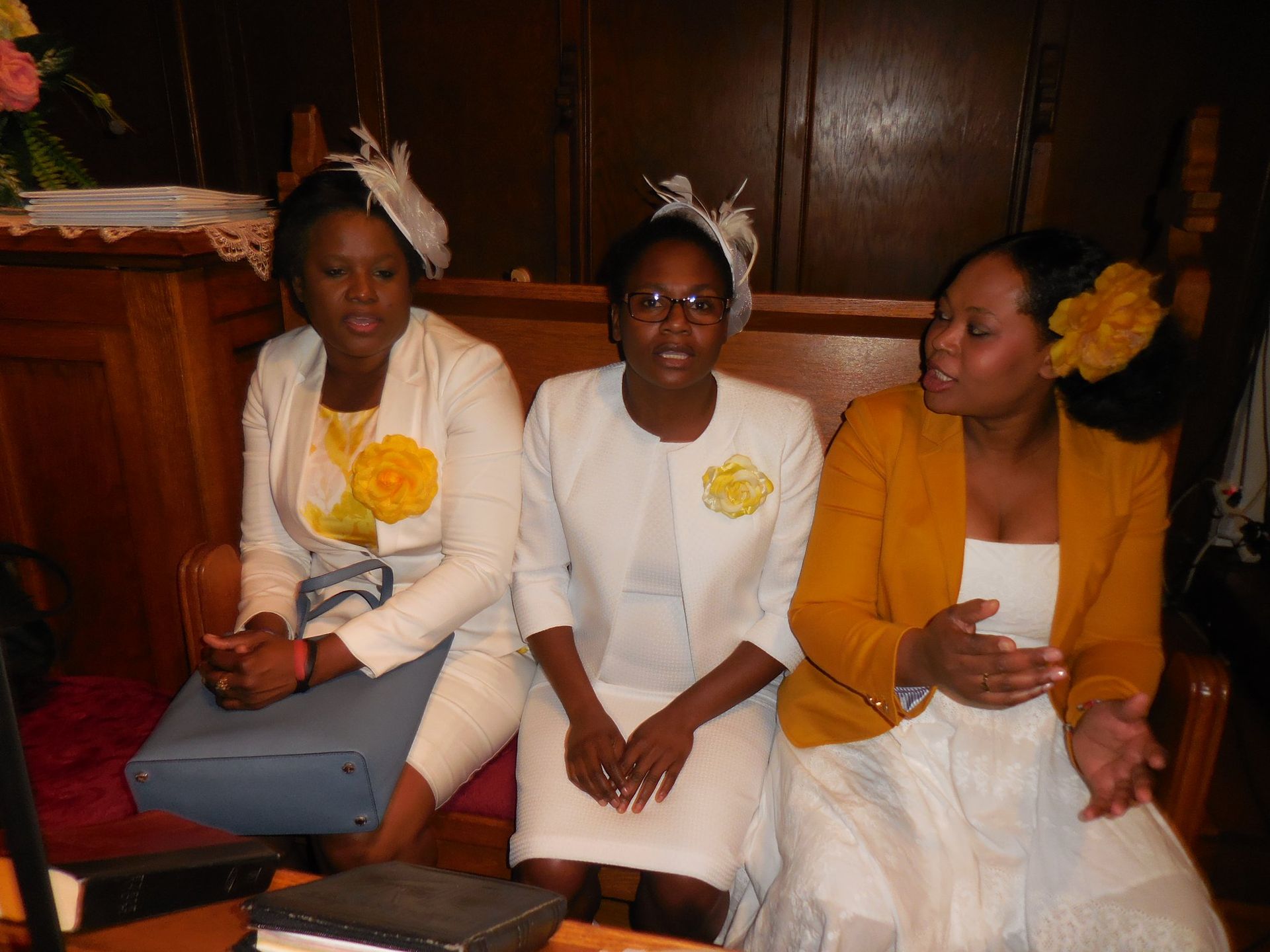Three people seated on wooden pews, wearing white and mustard yellow jackets with decorative floral headpieces.