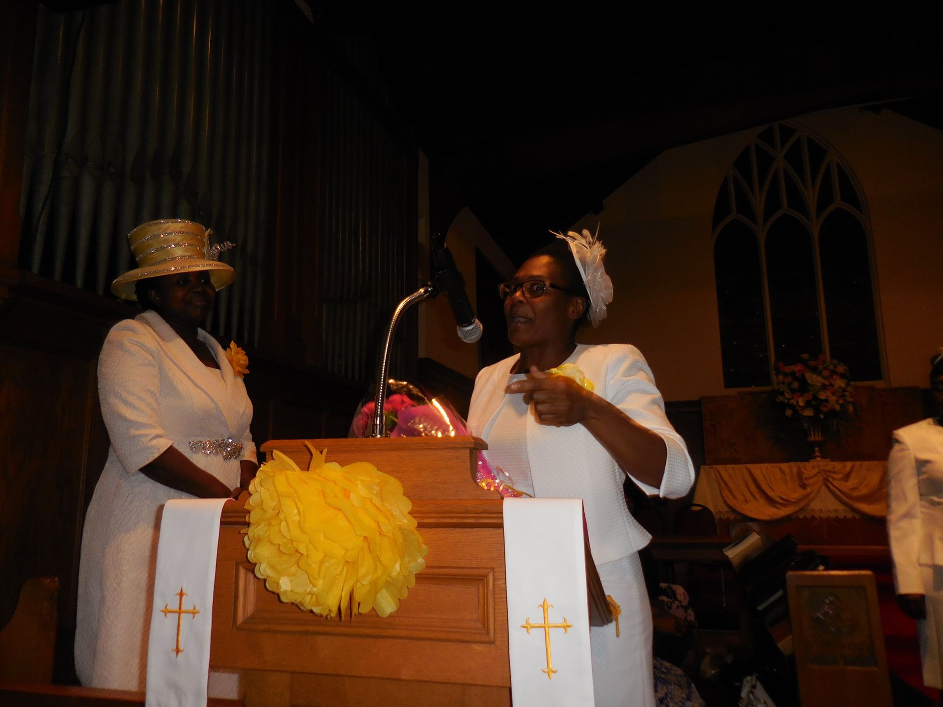 Two individuals in white clothing and hats stand at a church lectern, one speaking to an audience.