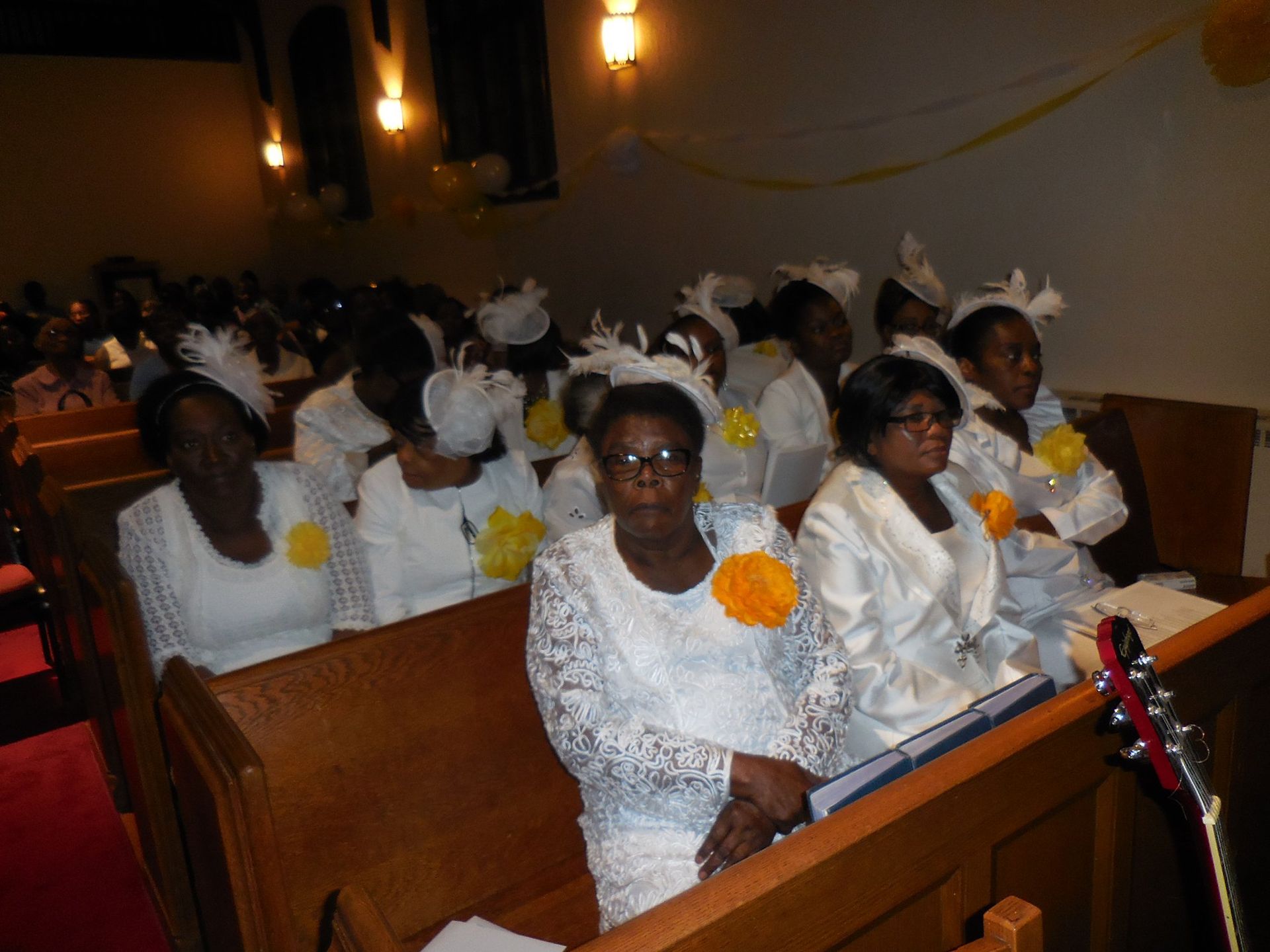 A group of people wearing white formal attire with yellow accents and hats seated in a wooden church pew.