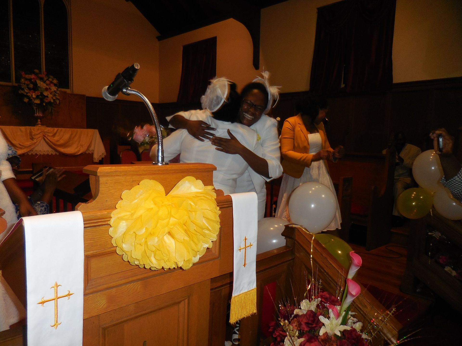 Two people in white attire hug joyfully behind a wooden church pulpit decorated with a yellow flower and white cloths.