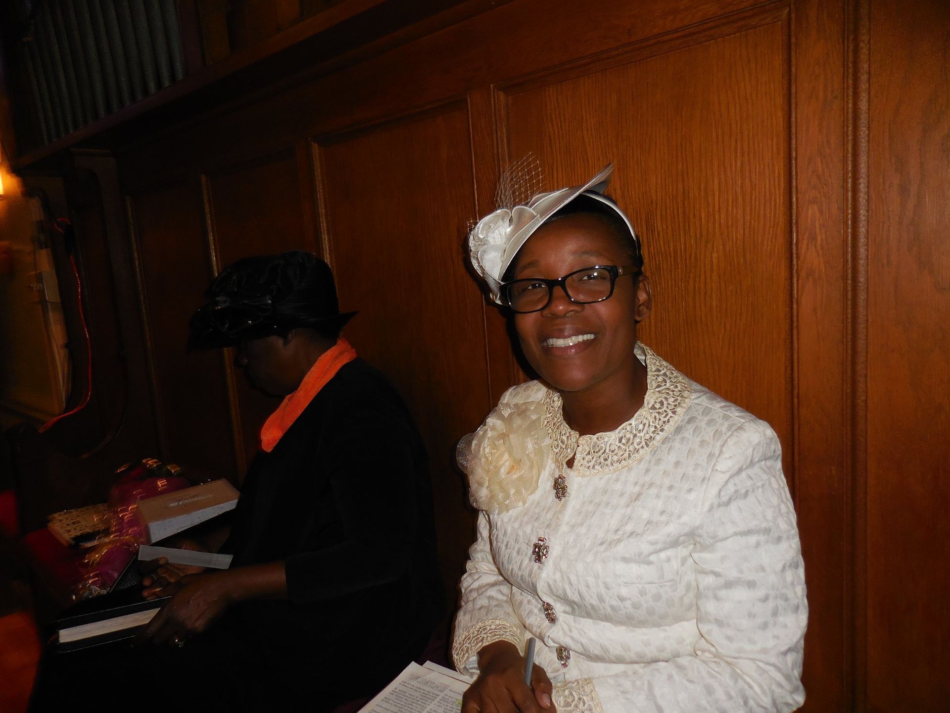 A person in a white textured suit and decorative hat smiles while sitting next to another person in a dark room.