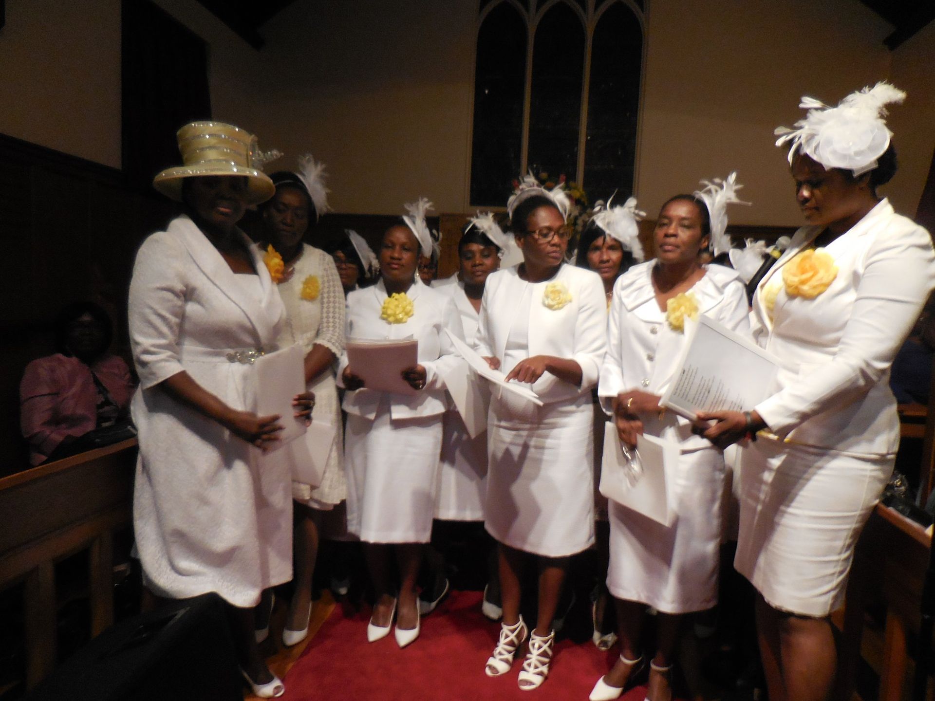 A group of people wearing matching white suits and hats stand in a church, holding programs and singing.