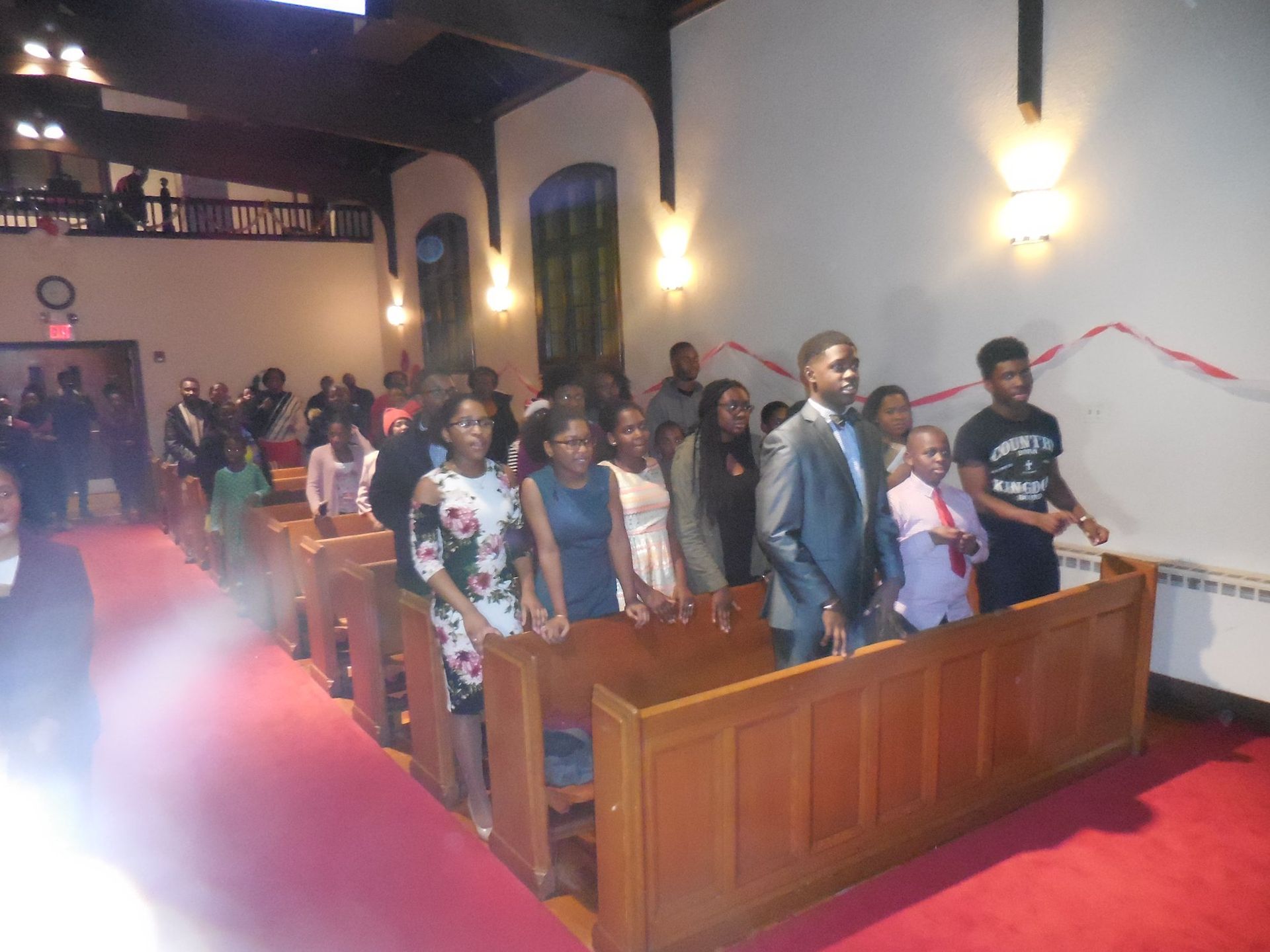 People stand in pews during a gathering inside a church with a red carpet aisle.
