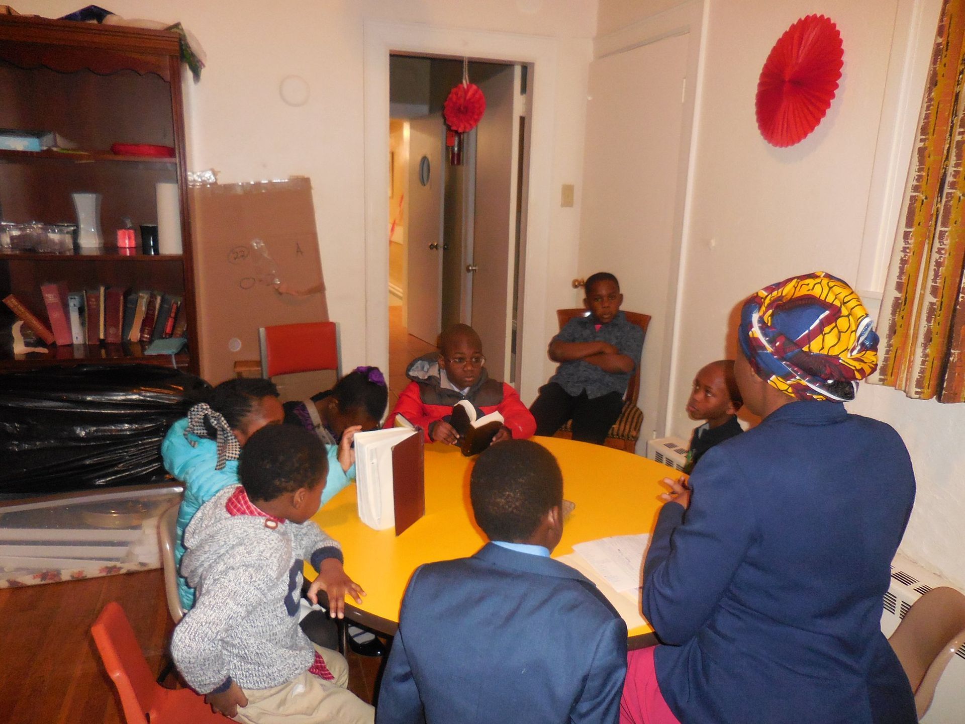 A group of people sit around a circular yellow table in a room, some reading books, while one person leads the session.