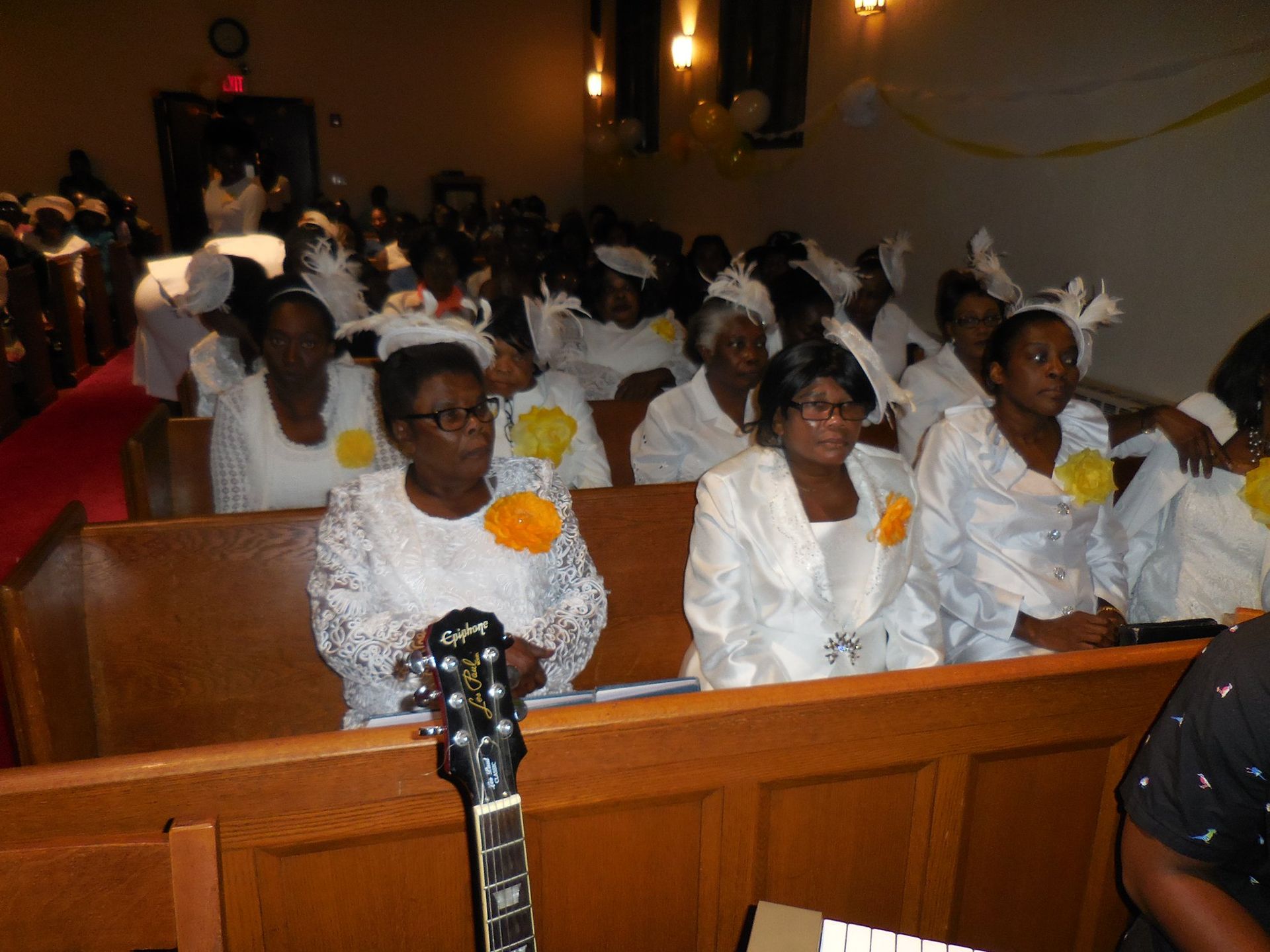 A congregation dressed in white seated in wooden church pews, with a guitar neck in the foreground.