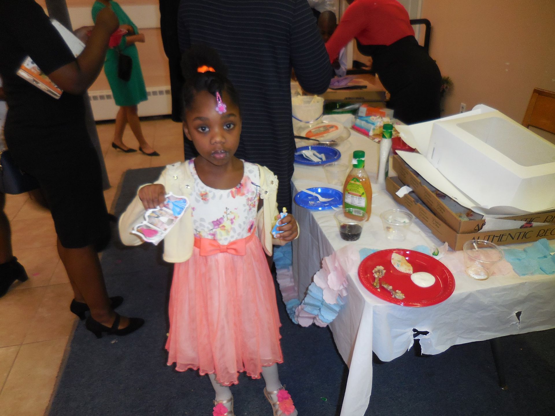 A child stands holding a small toy in front of a table laden with food and serving supplies at an indoor gathering.