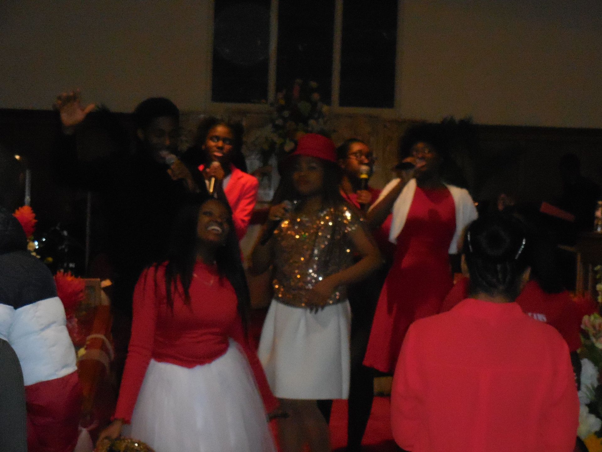 A group of people singing on stage, wearing red and white outfits, performing in a dim indoor setting.