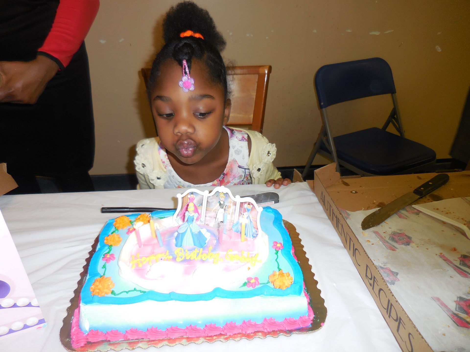 A child leans in to blow out candles on a rectangular cake decorated with cartoon characters on a white tablecloth.