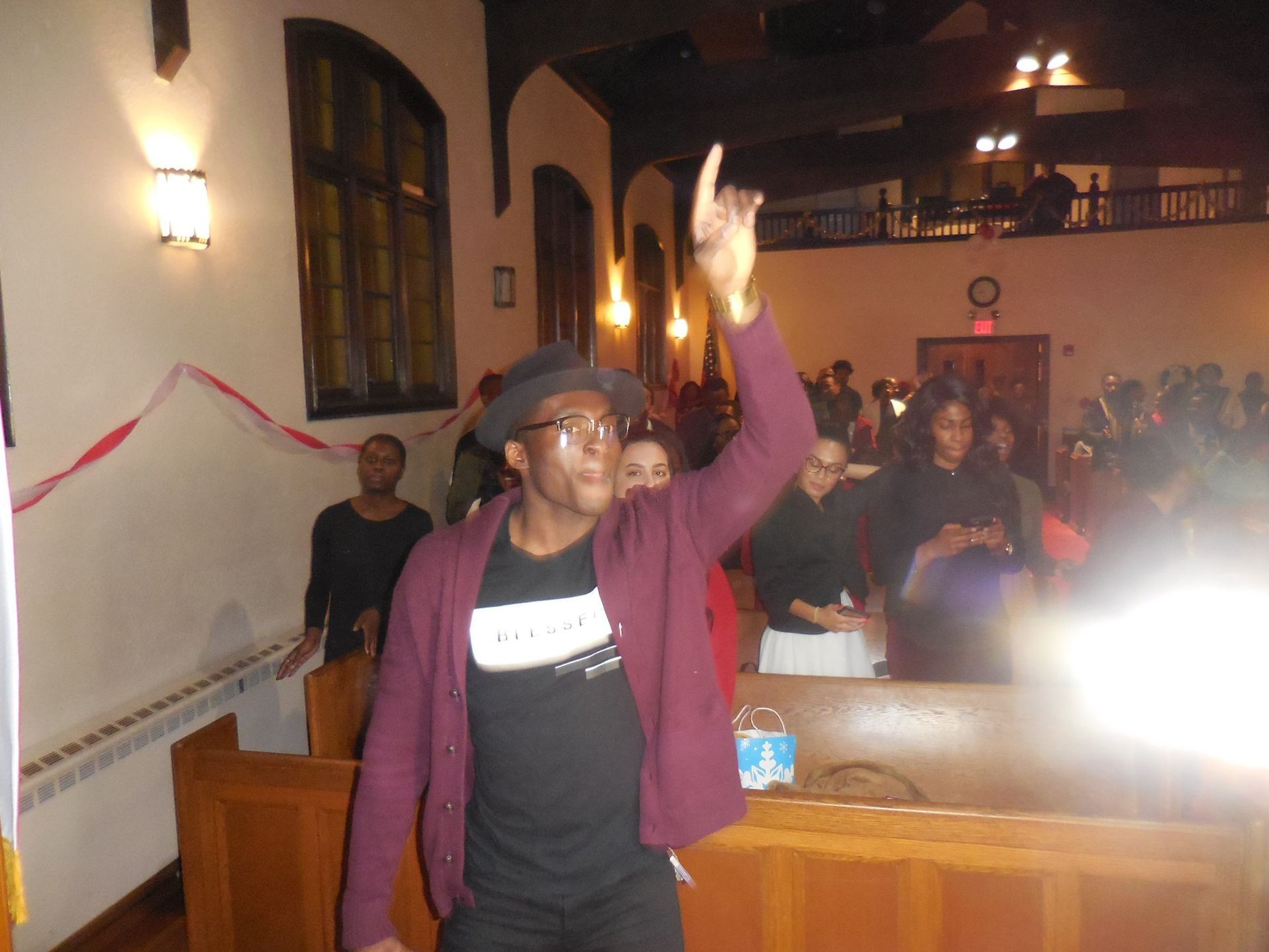A person in a hat and maroon cardigan stands in a church with one hand raised, while others sit in pews behind them.