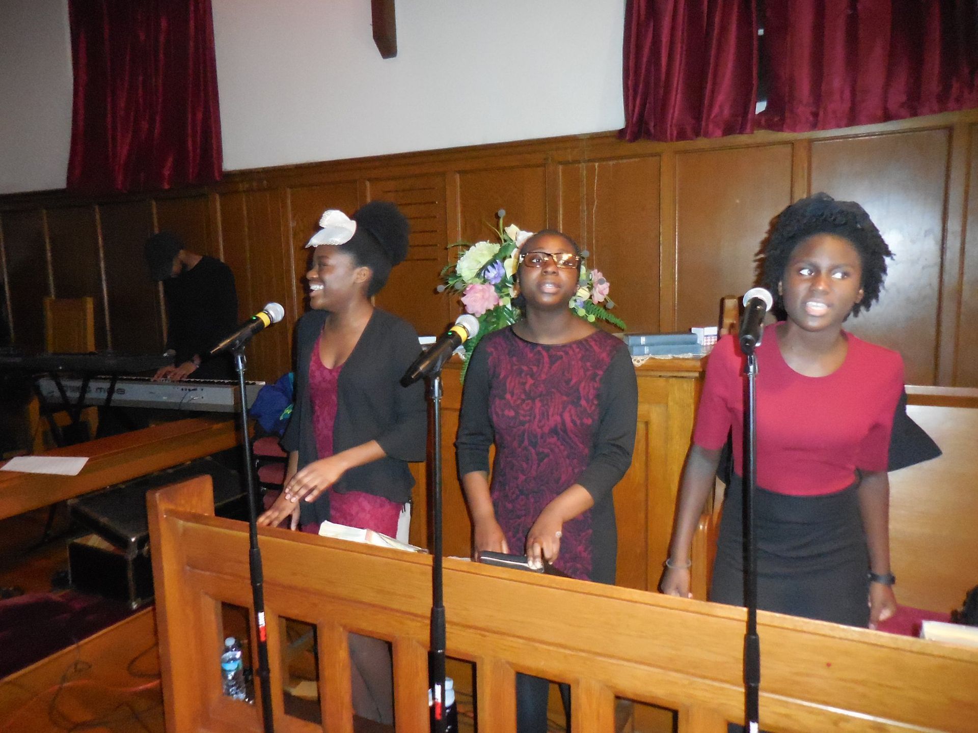 Three singers perform at microphones in front of wooden paneling and red curtains in a church setting.