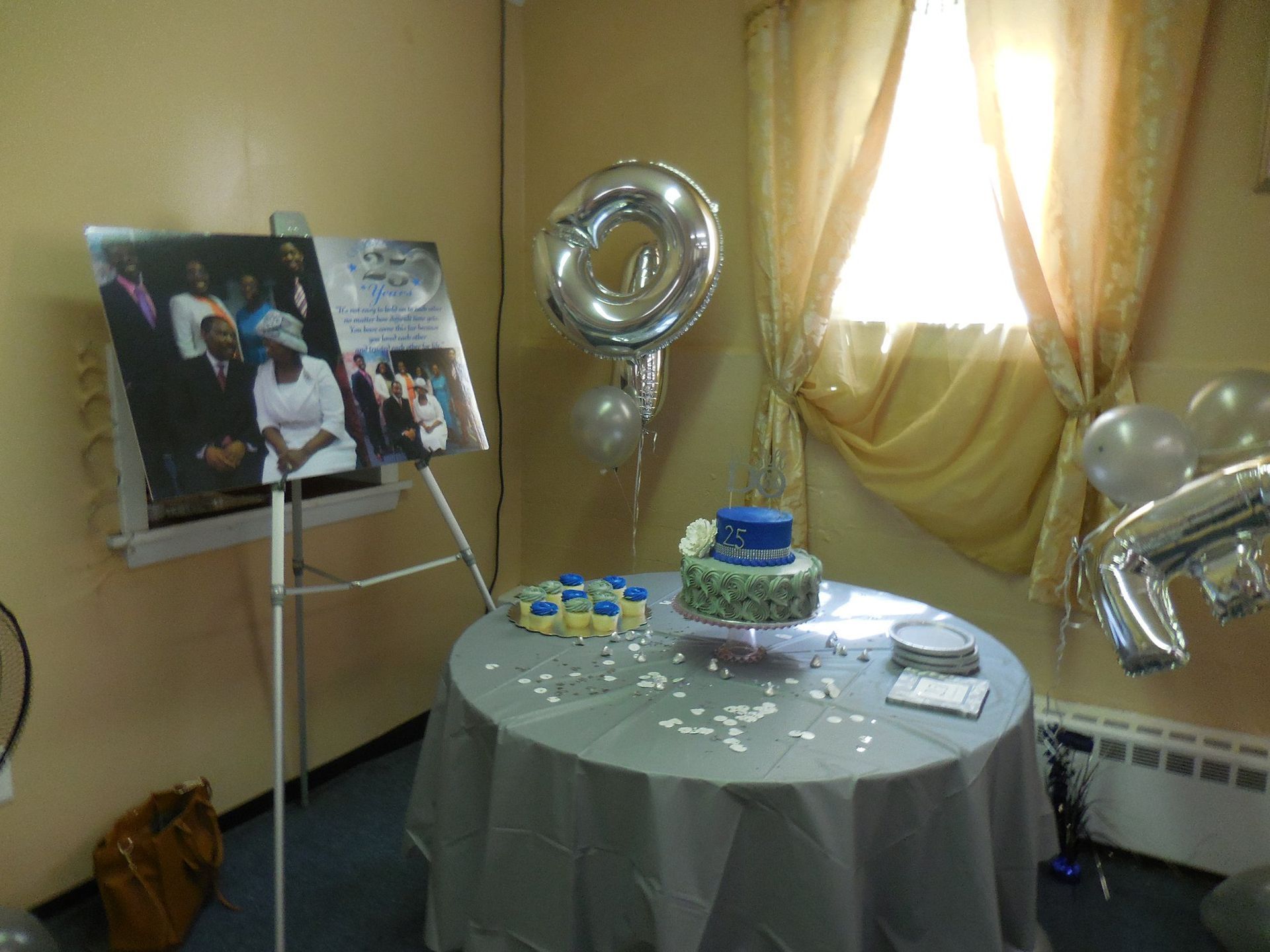 A celebratory display featuring a photo collage on an easel, a tiered cake on a draped table, and silver number balloons.