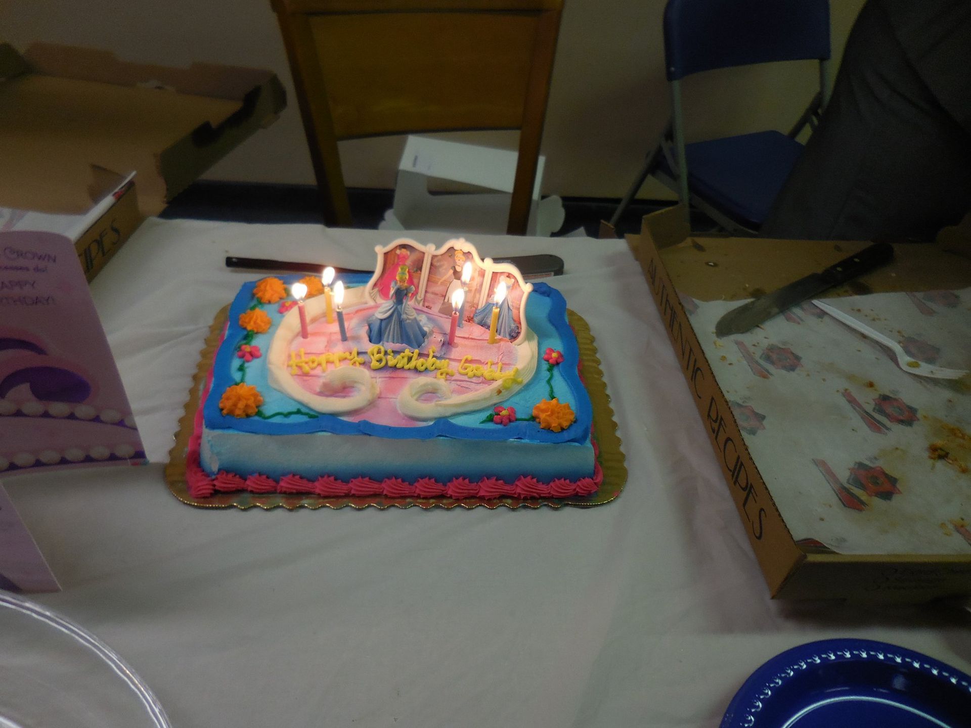 A rectangular birthday cake with lighted candles sits on a table, next to an open pizza box and a birthday card.
