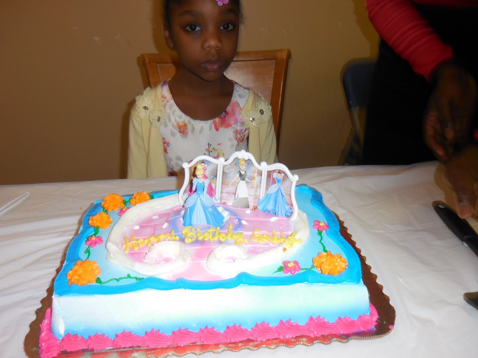 A child sits behind a rectangular cake decorated with blue, pink, and orange icing and Cinderella cake toppers.