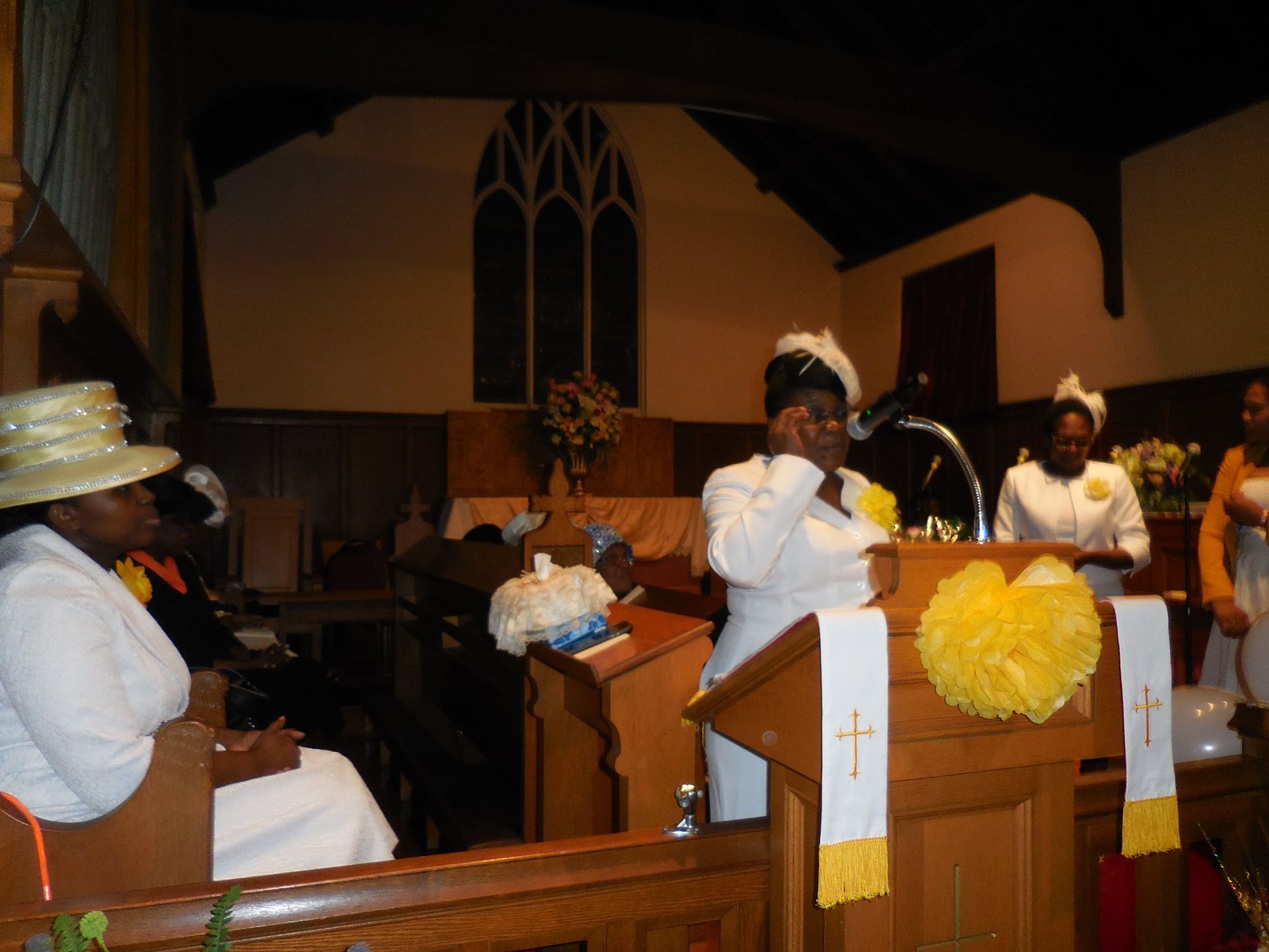 A woman stands at a wooden pulpit in a church, speaking into a microphone, while others sit nearby in white attire.