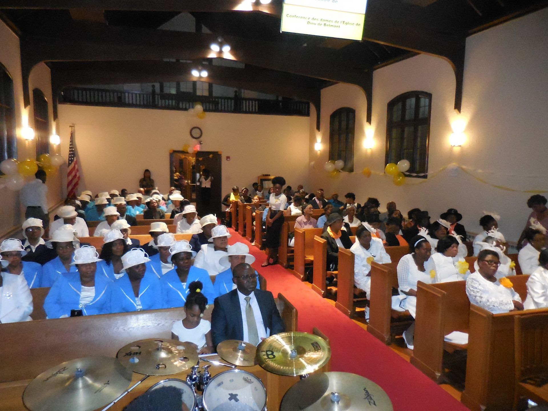 Congregation in formal white attire gathered in a church for a service, viewed from behind the musical instruments.