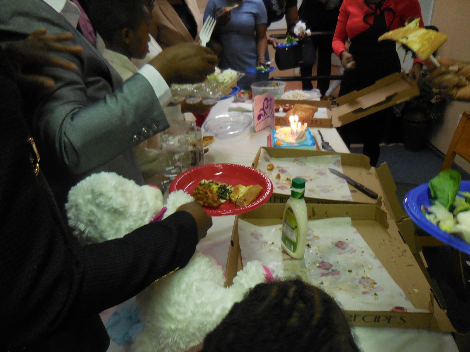 A celebration with a small lit cake, pizza boxes, and food on plates, with hands gathering around a table.