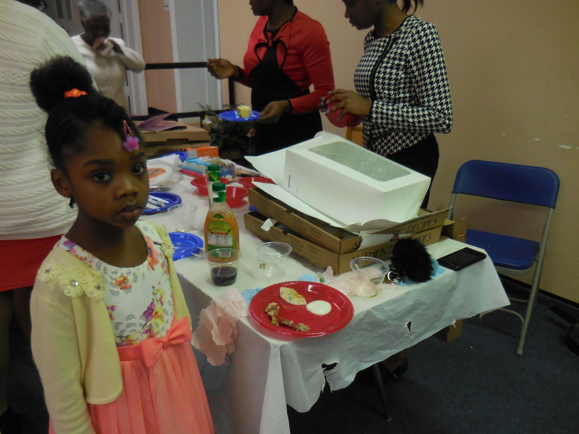 A child in a light-colored dress stands by a table with pizza boxes and snacks at an indoor gathering.