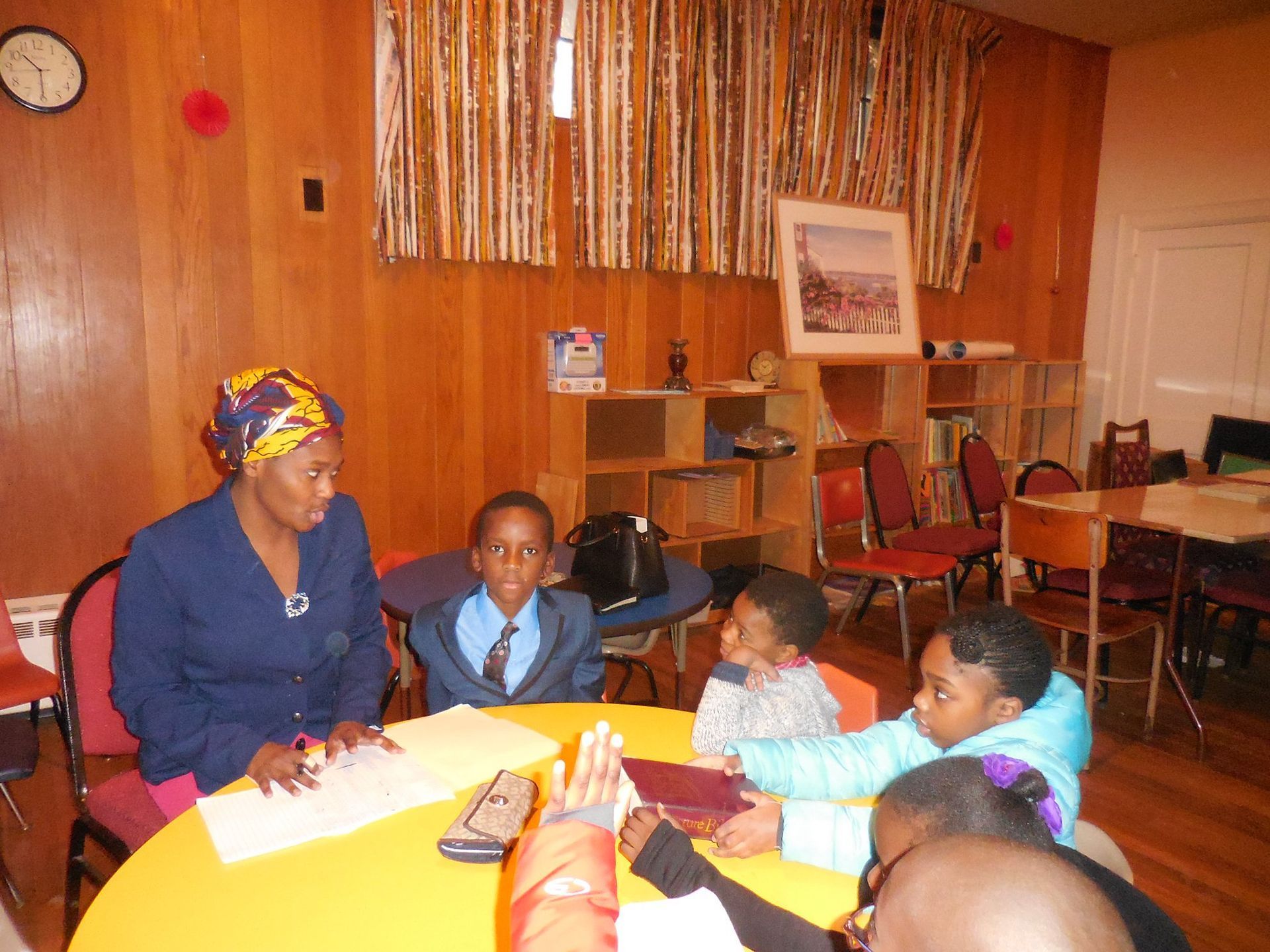 A woman sits at a round yellow table with a group of children in a wood-paneled room, participating in a group activity.