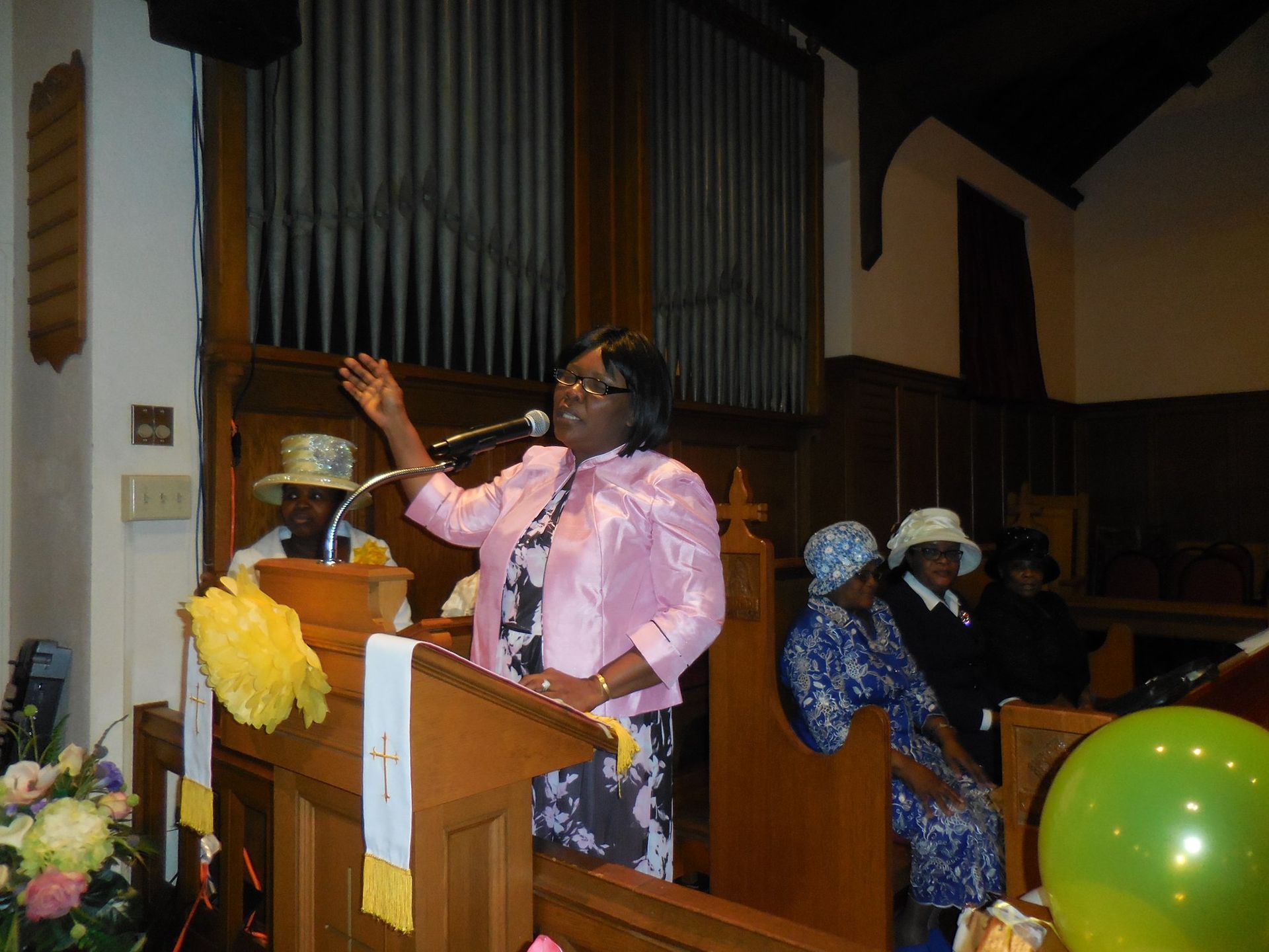 A person in a pink jacket speaks at a wooden podium in a church, with attendees seated nearby and a green balloon.