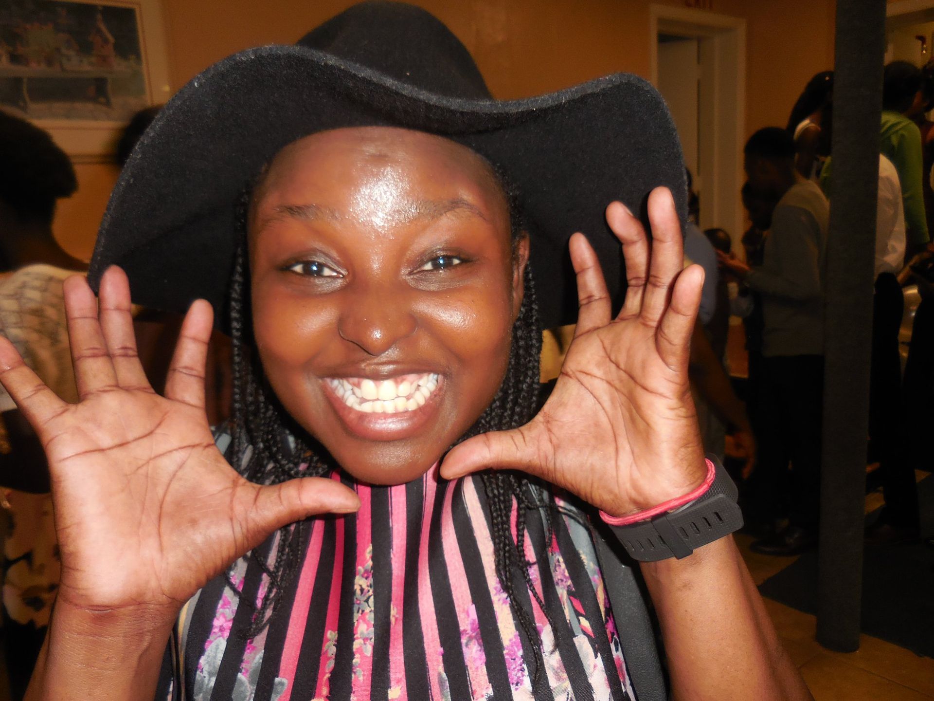 A person wearing a wide-brimmed black hat and striped top, smiling while framing their face with their open hands.