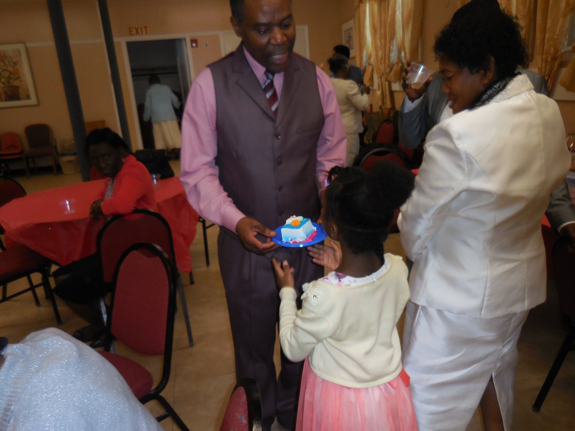 A person in a vest holds a small cake on a blue plate for a child in a pink skirt while guests socialize in a hall.