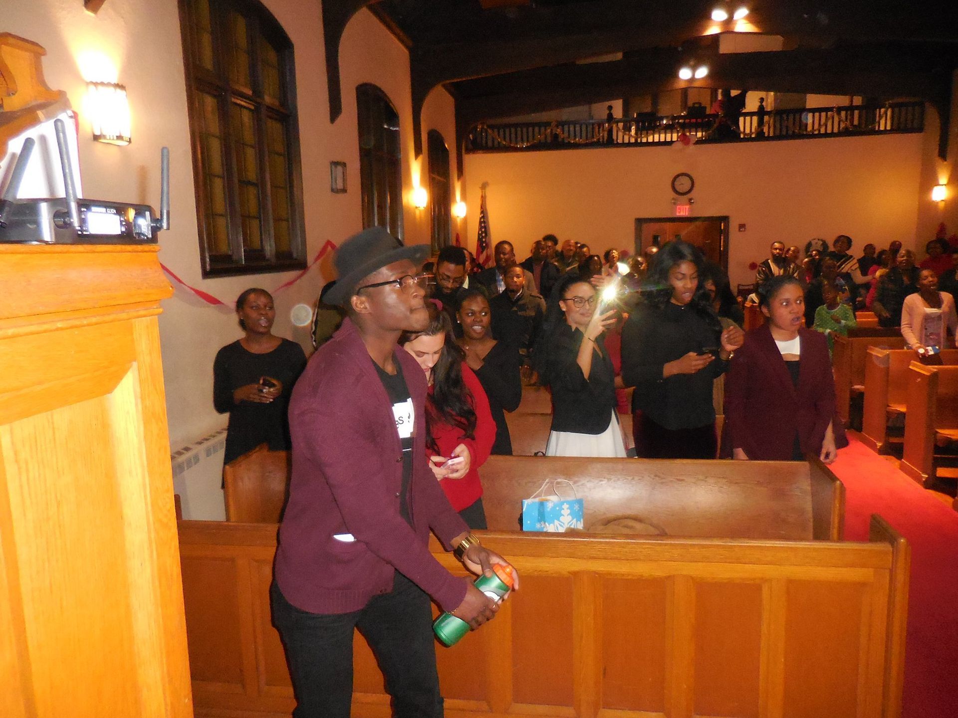 A young person in a dark hat and sweater holds a canister in a church while others watch from the pews.