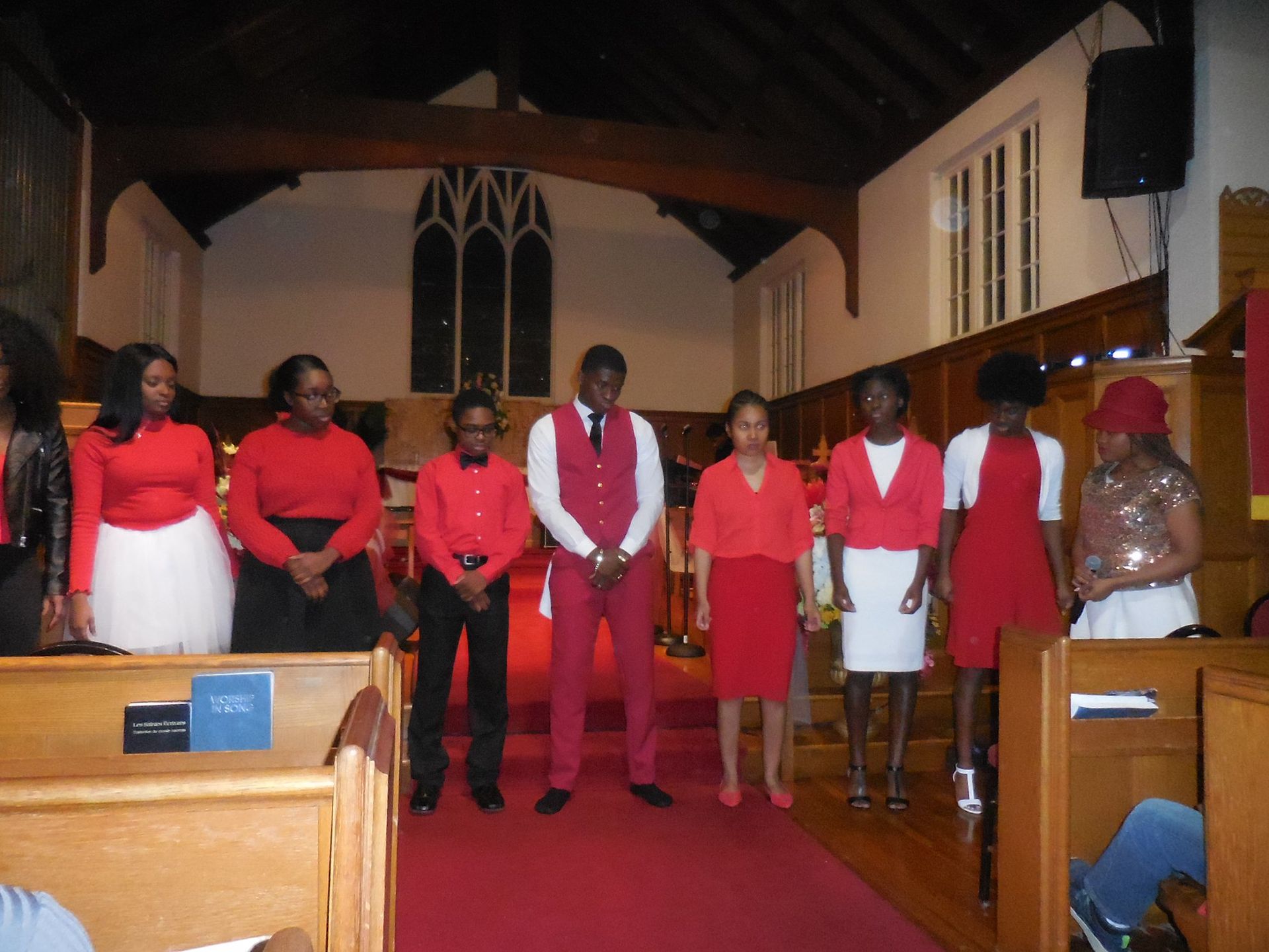 A group stands in a church aisle, wearing red, white, and black formal attire, facing forward in a performance setting.