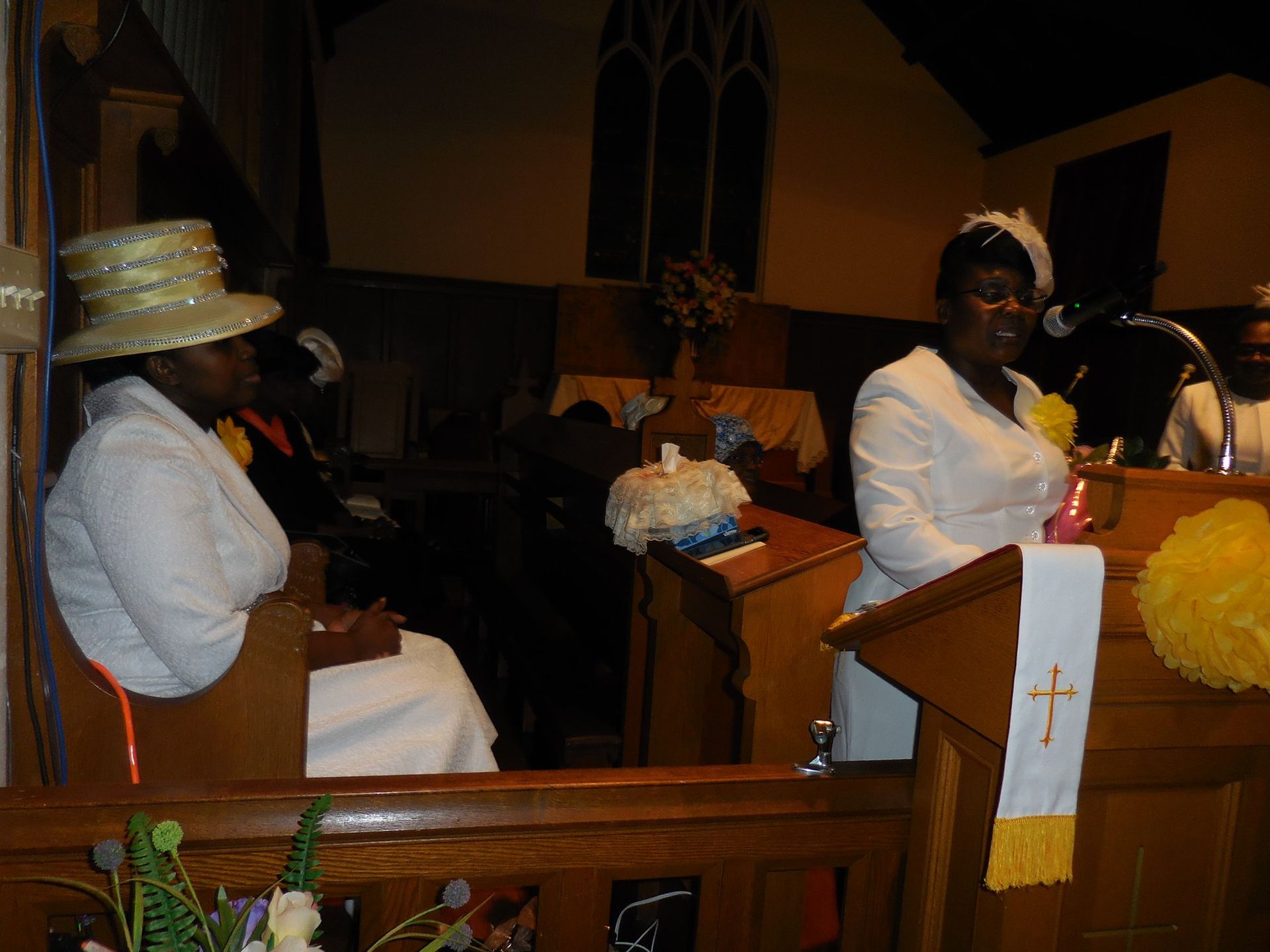 Two people in formal white attire and hats attend a church service, with one speaking from a wooden pulpit.