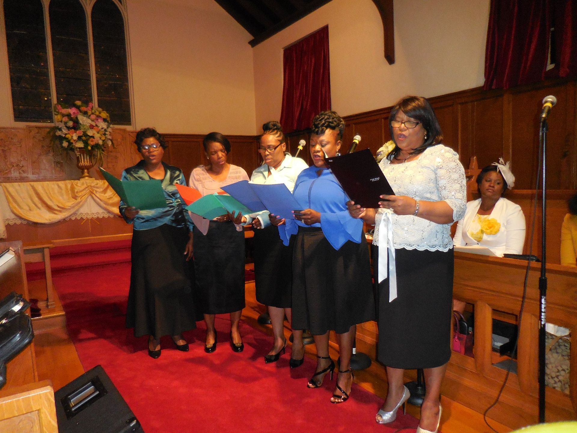 A group of people standing in a row in a church, singing from folders held in their hands.