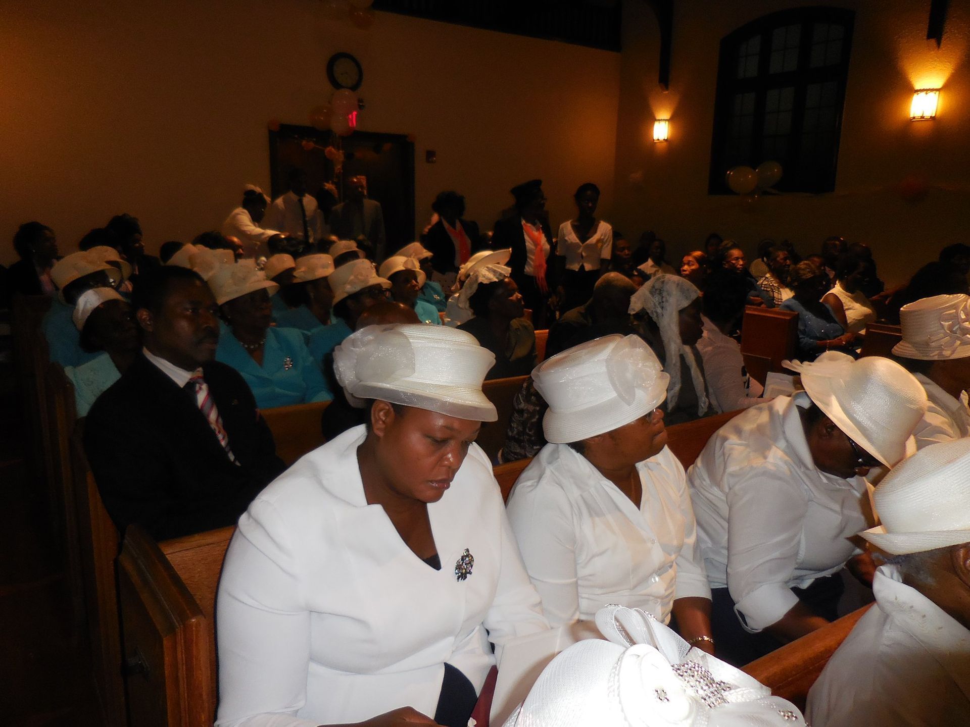 People in white hats and attire sit in church pews during a service in a dimly lit, wood-paneled building.