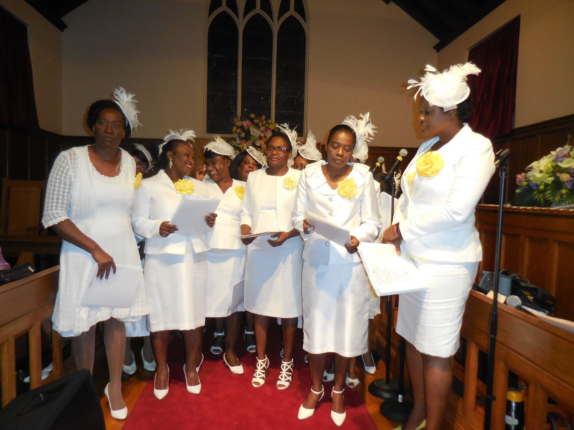 A group of people wearing white suits and hats stand in a church, holding papers and appearing to sing together.