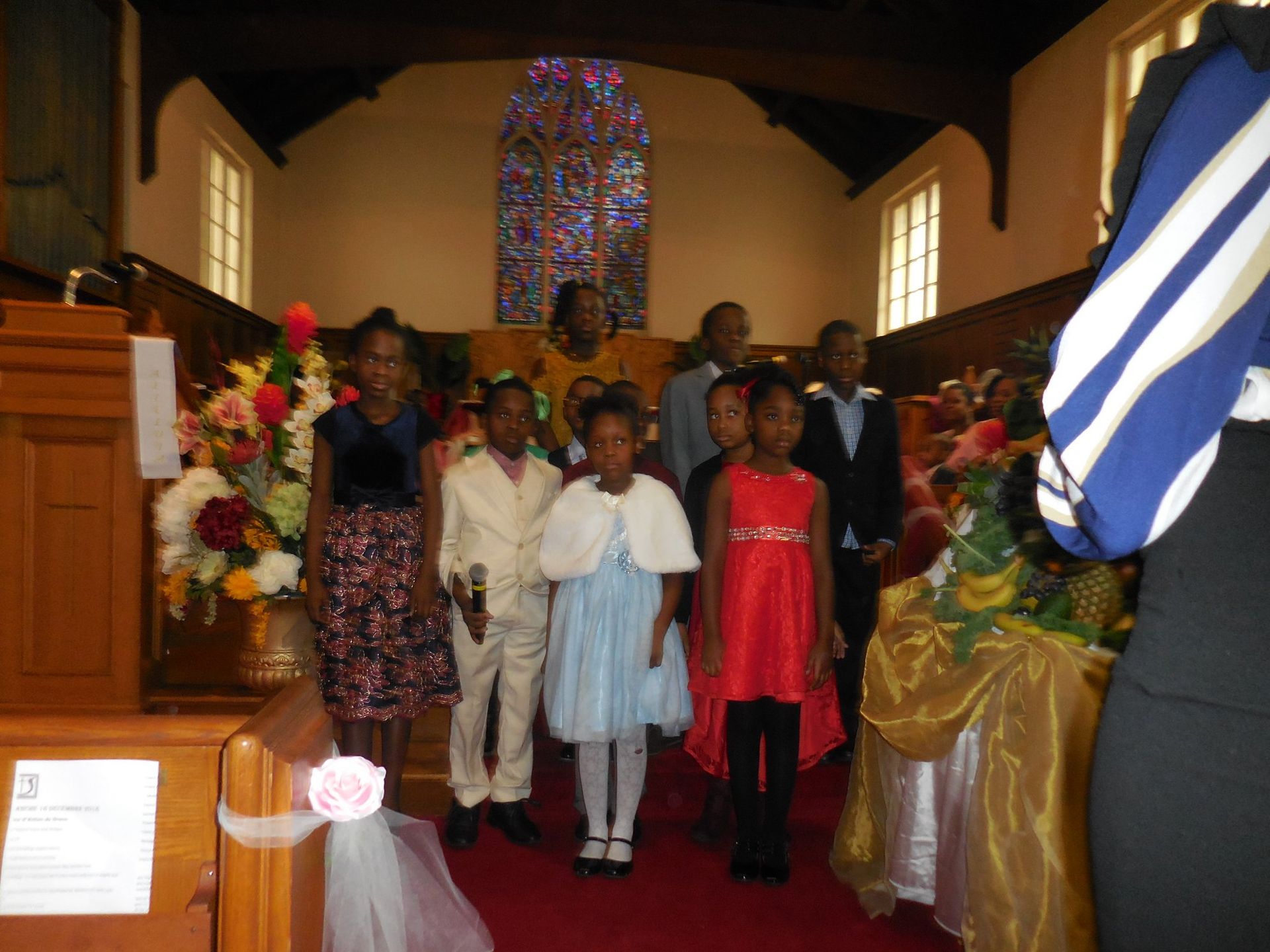 A group of children in formal attire stands on a red carpet in a church, with a stained-glass window in the background.