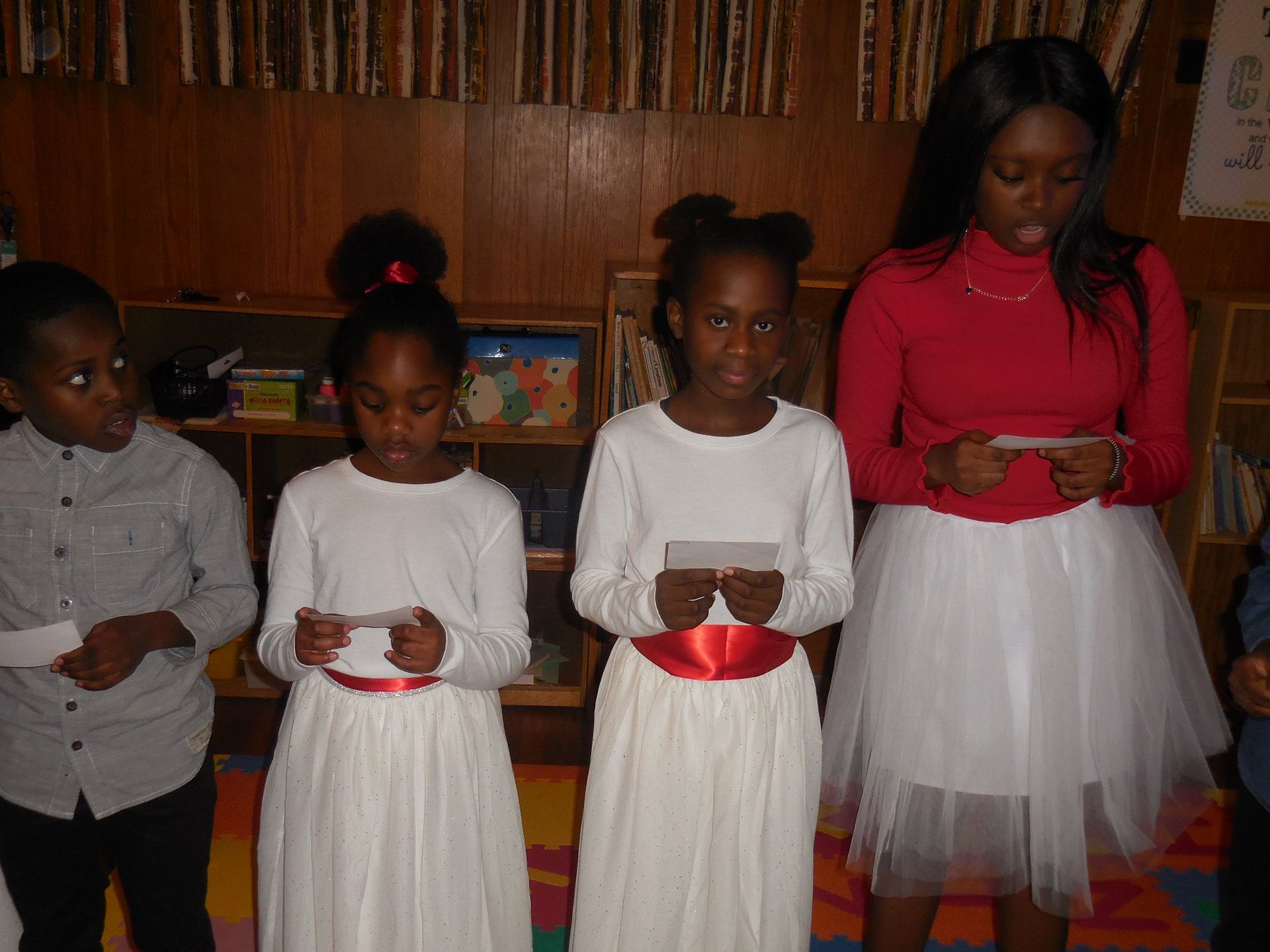 Four people stand in a line against a wood-paneled wall, each holding a small piece of paper while looking down at them.