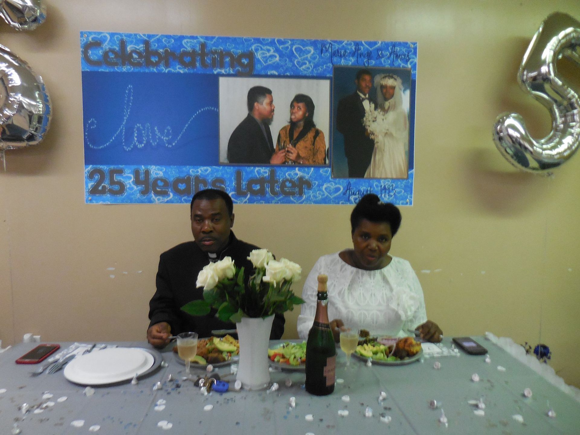 A couple sits at a table celebrating their 25th anniversary, with a photo banner above them and party decor.