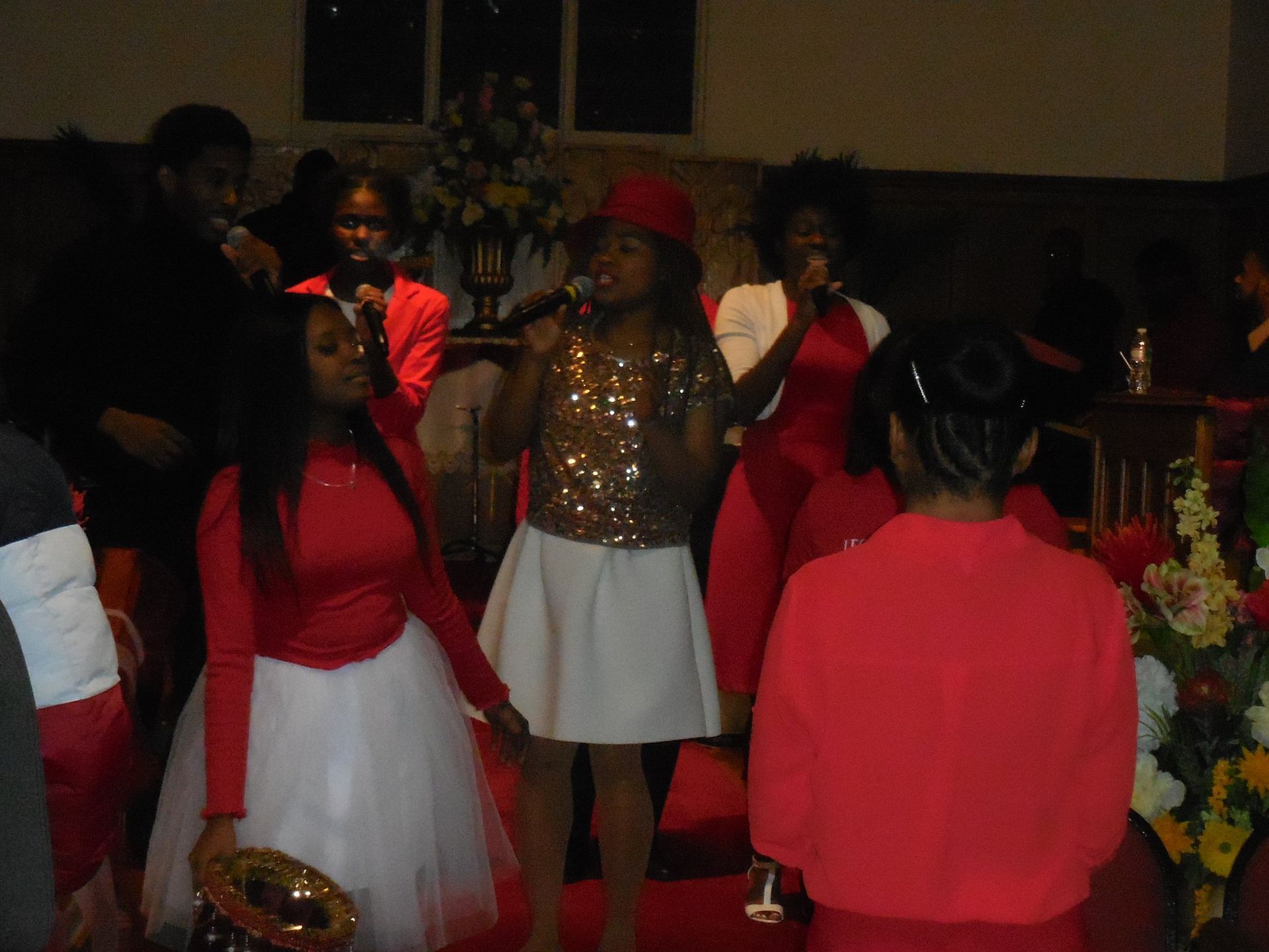 A group of people wearing red and white clothing perform in a church, singing into microphones during a musical service.