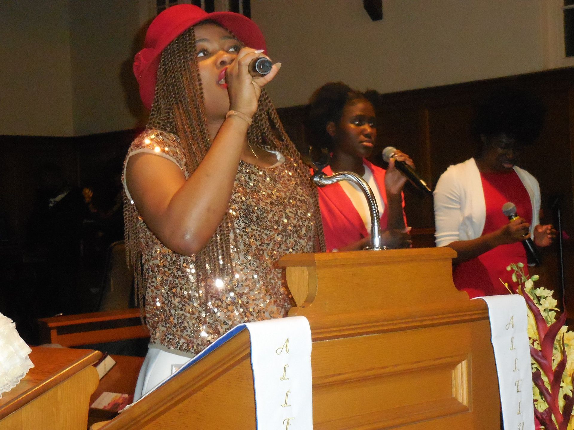 Three people stand behind a wooden pulpit, singing into microphones in a dimly lit indoor setting.