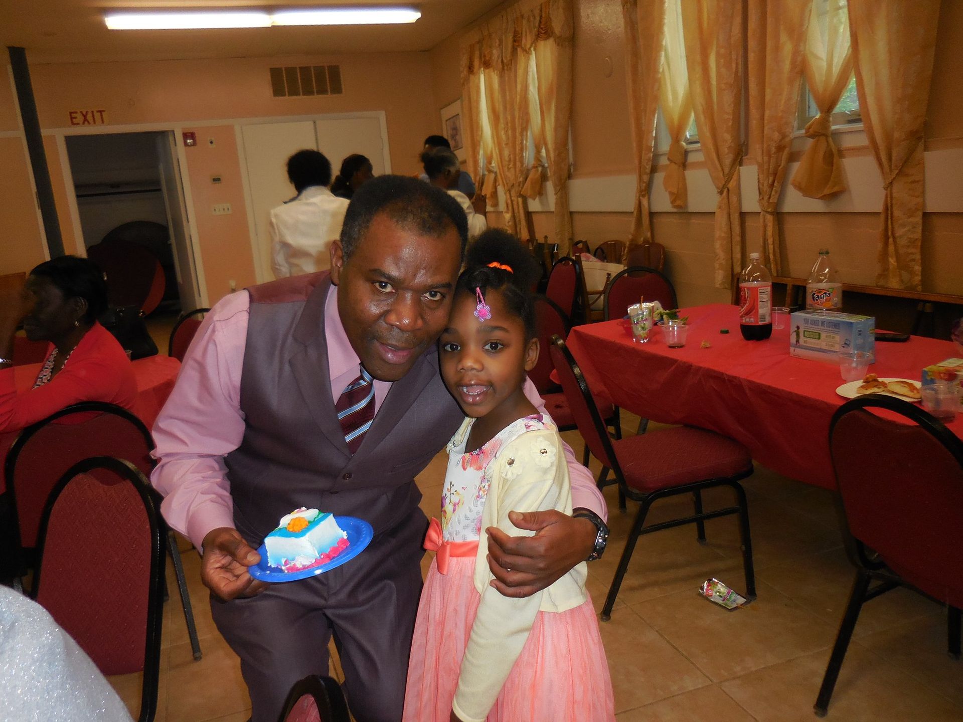A smiling man in a vest holds a small cake, standing with a girl in a pink dress at a festive gathering.