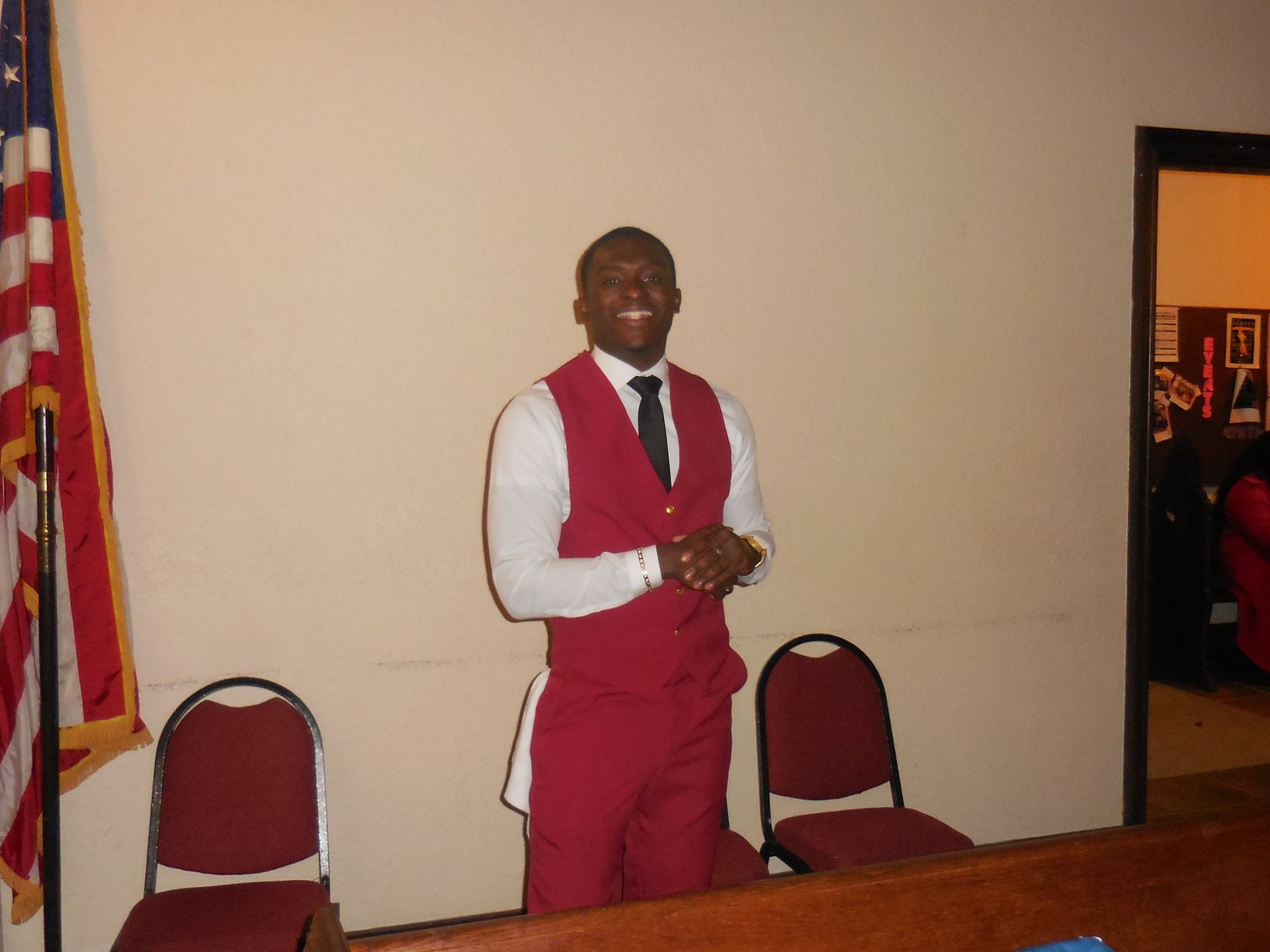A smiling person wears a maroon vest, white shirt, and black tie, standing in a room with an American flag and two chairs.