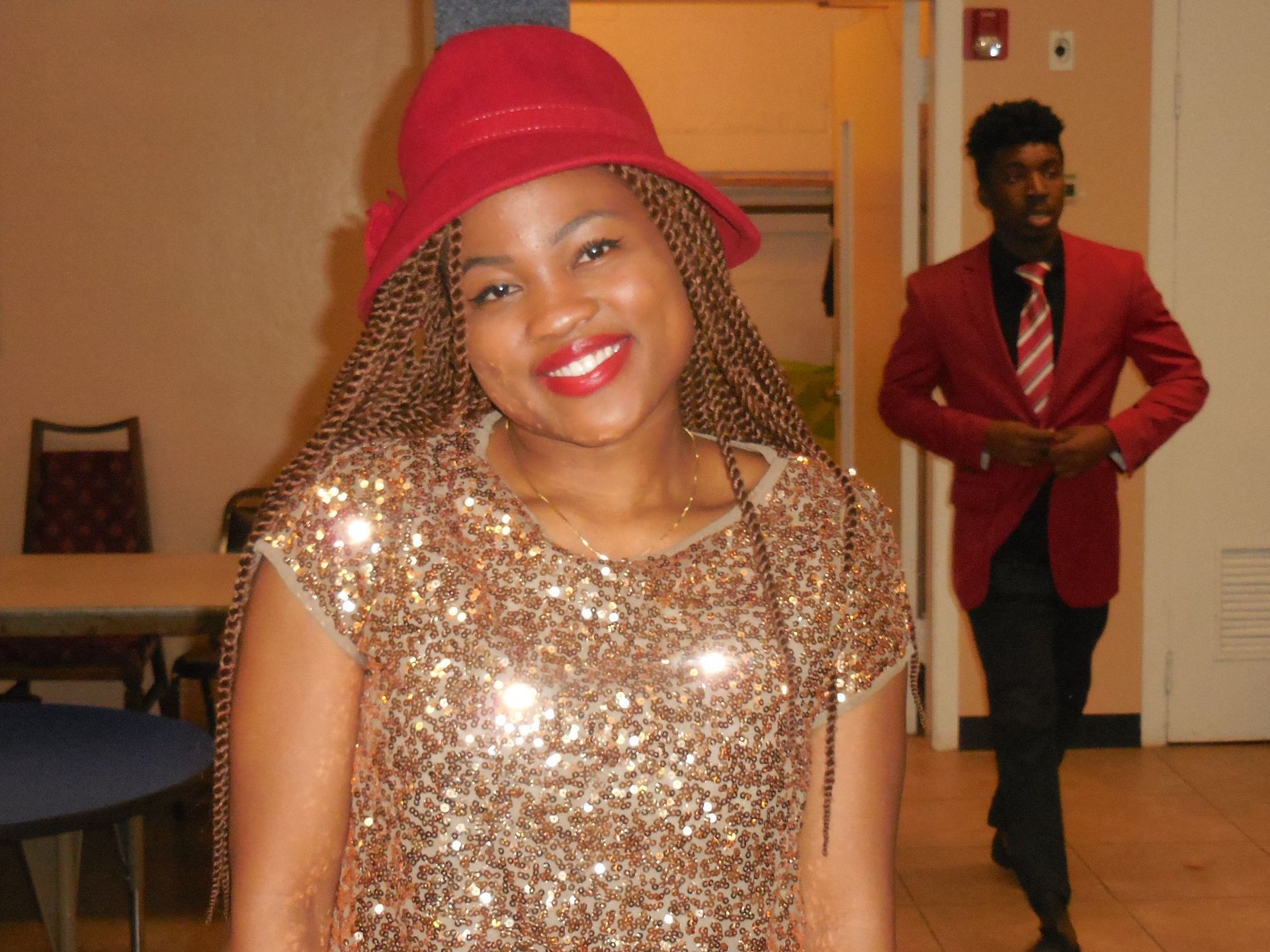 A smiling person in a red hat and sequined top with a person in a red blazer in the blurred background.