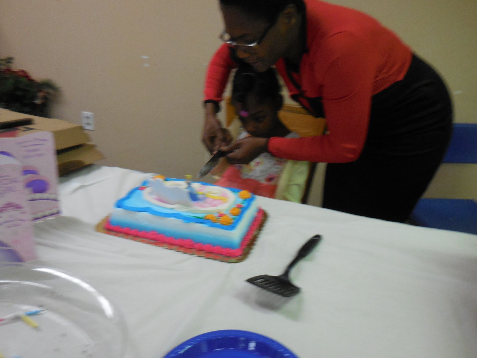 An adult assists a child in cutting a rectangular birthday cake decorated with blue frosting on a table.
