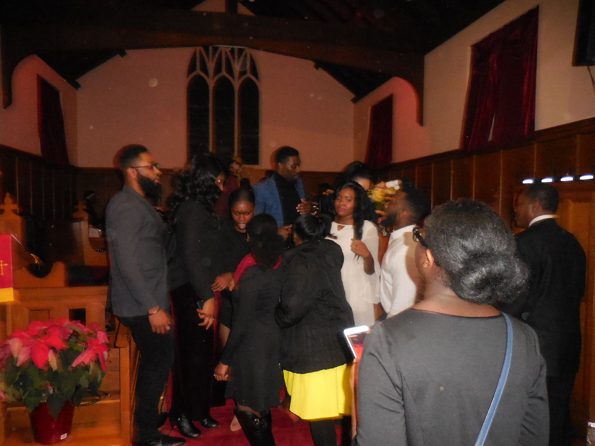 A group of people gathers in a wood-paneled church, some standing and talking near a red-clothed pulpit with poinsettias.