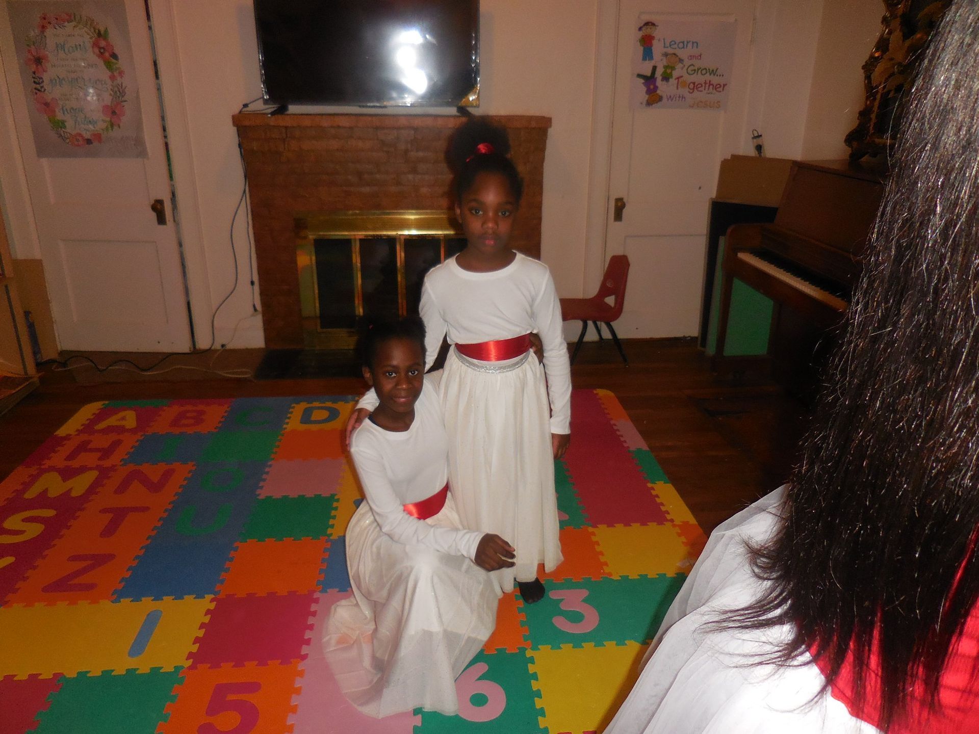 Two individuals in long white dresses with red waistbands stand and kneel on a colorful, numbered foam play mat.