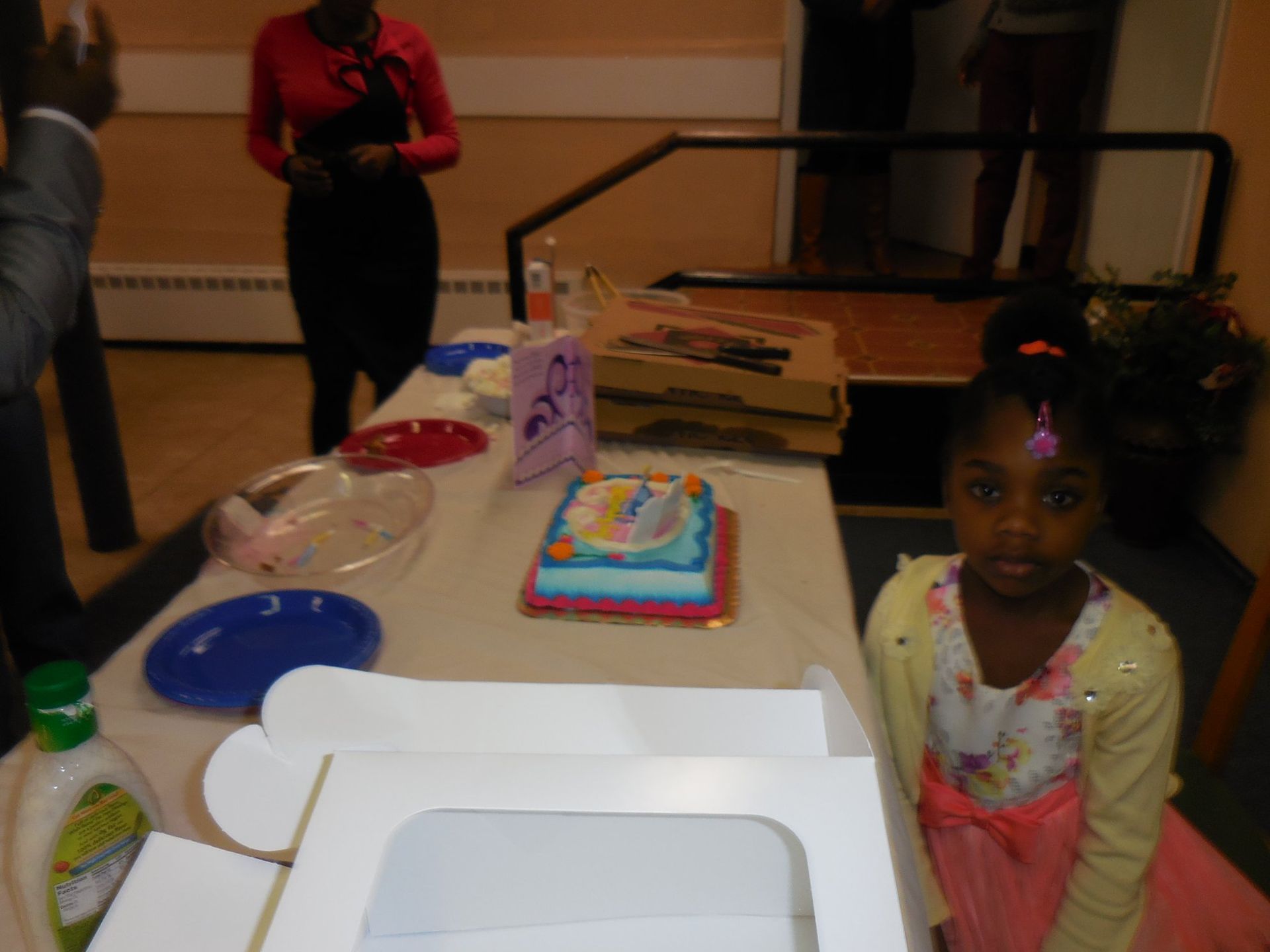 A child stands by a table set for a celebration, featuring a decorated cake, plates, and pizza boxes in a room.