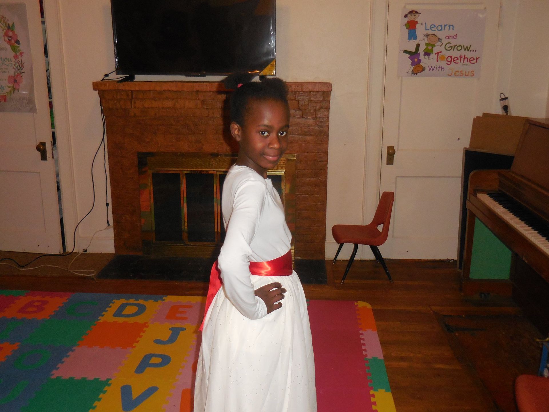 A person wearing a white long-sleeved dress with a red sash poses with hands on hips in a room with a fireplace.