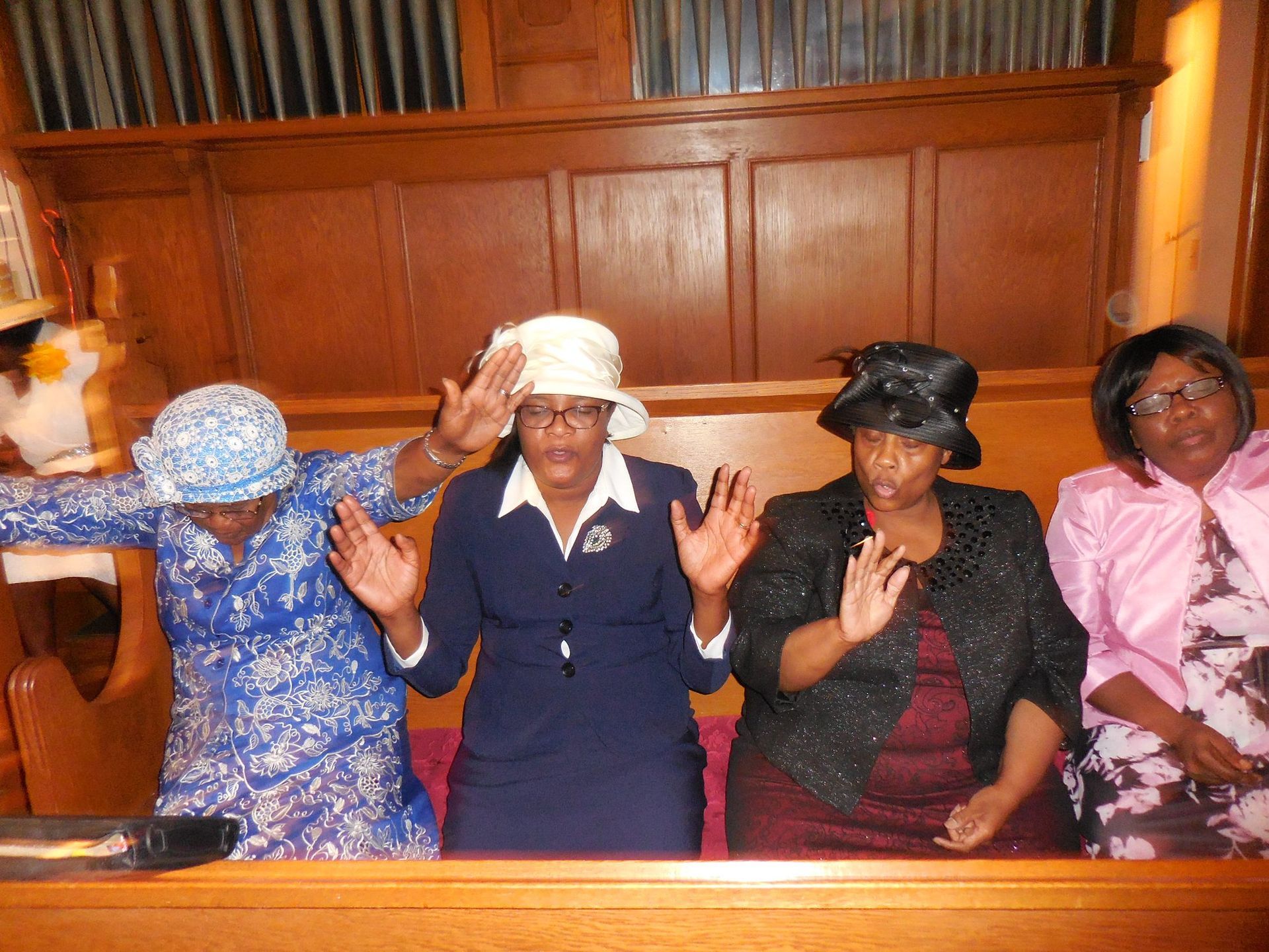 Four people in hats and formal attire sitting in a church pew with arms raised in prayer.